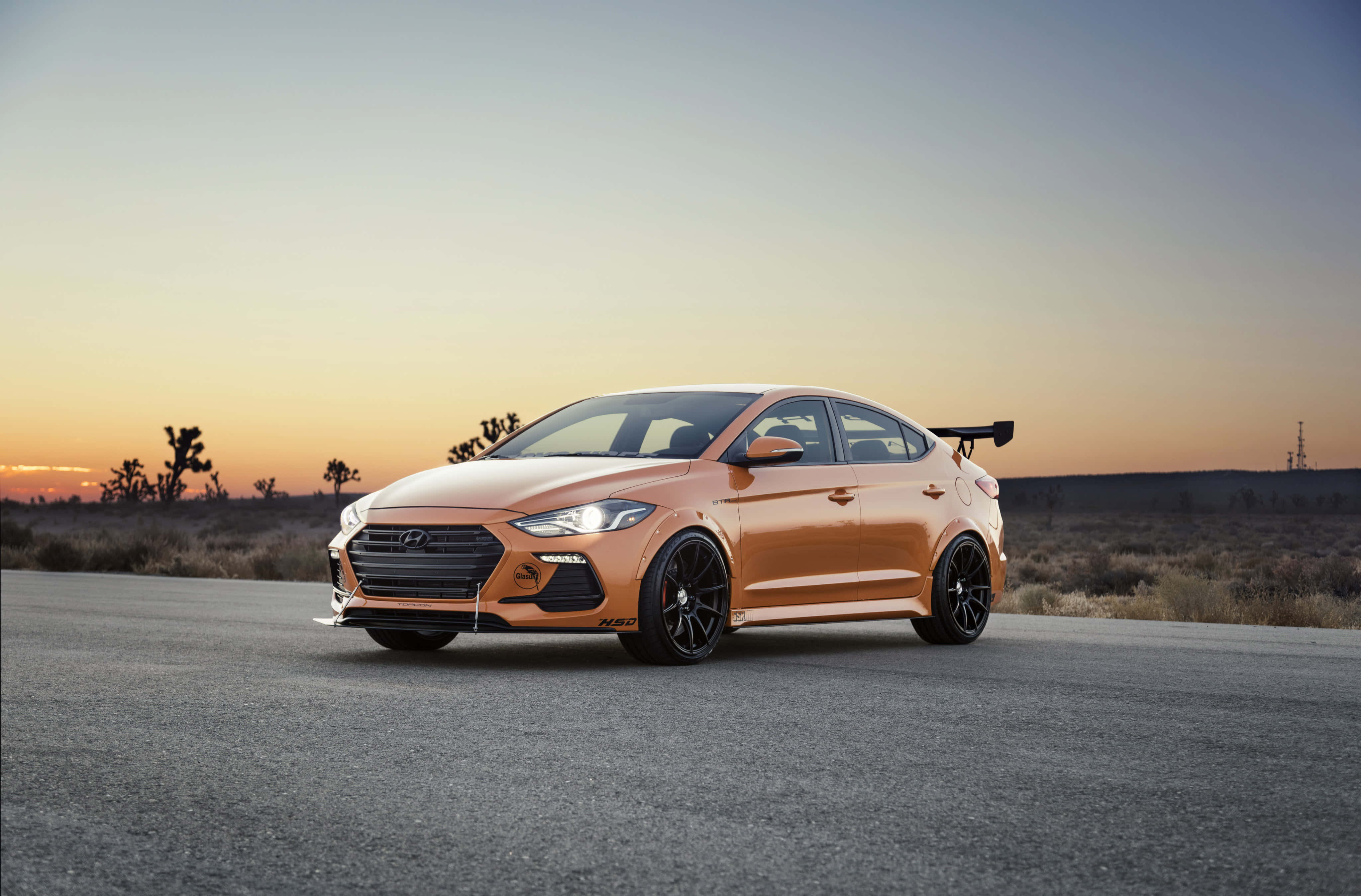 Hyundai Elantra 桌布 壁紙 — 一輛流線型的橙色Hyundai Elantra N在日落時分停在沙漠公路上，溫暖的金色光芒沐浴著其運動感十足的輪廓。