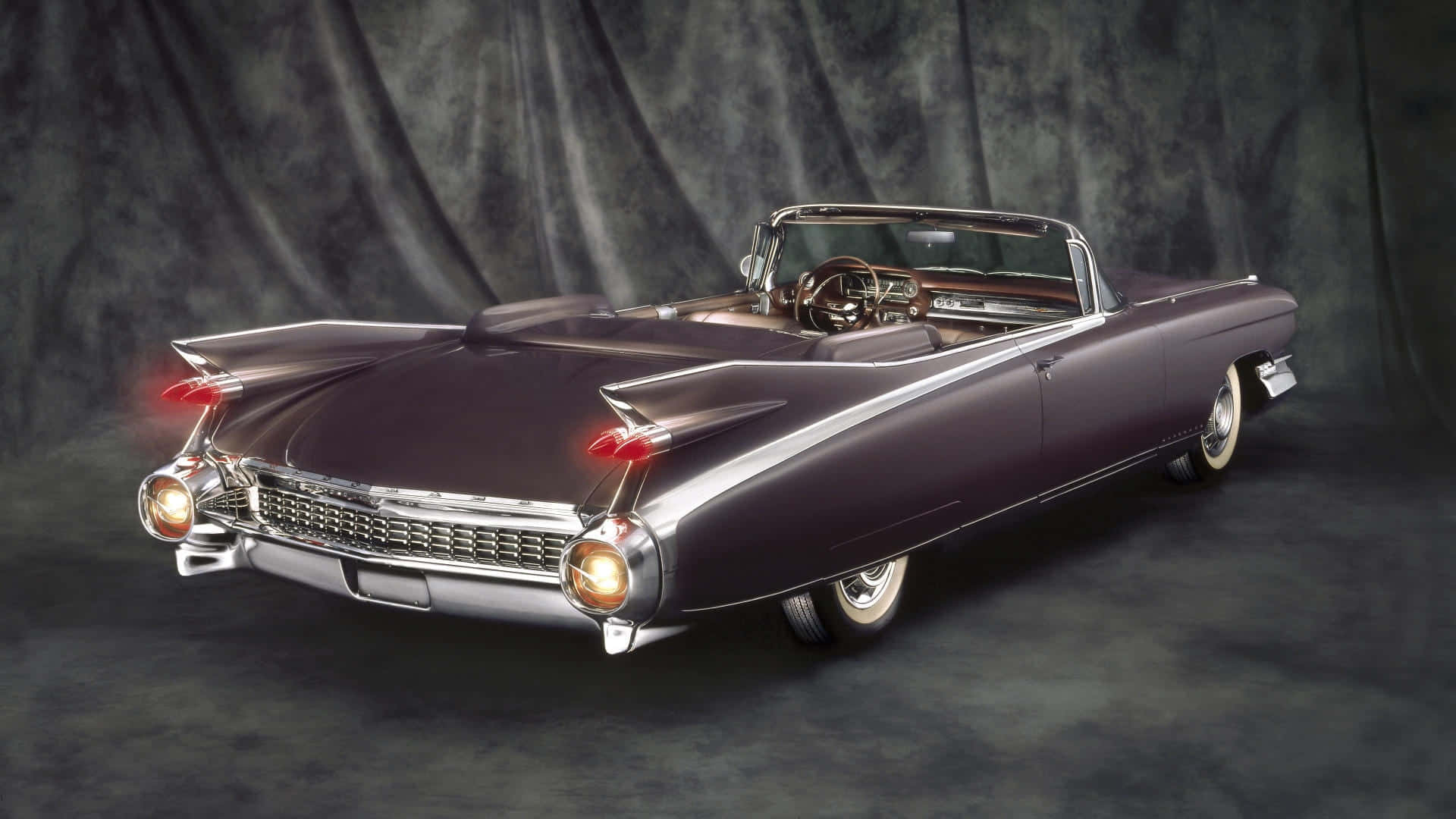 Cadillac Eldorado 桌布 壁紙 — 一輛時尚的紫色1950年代凱迪拉克敞篷車，帶有鍍鉻裝飾和發光的尾燈，停在深色垂簾背景前。