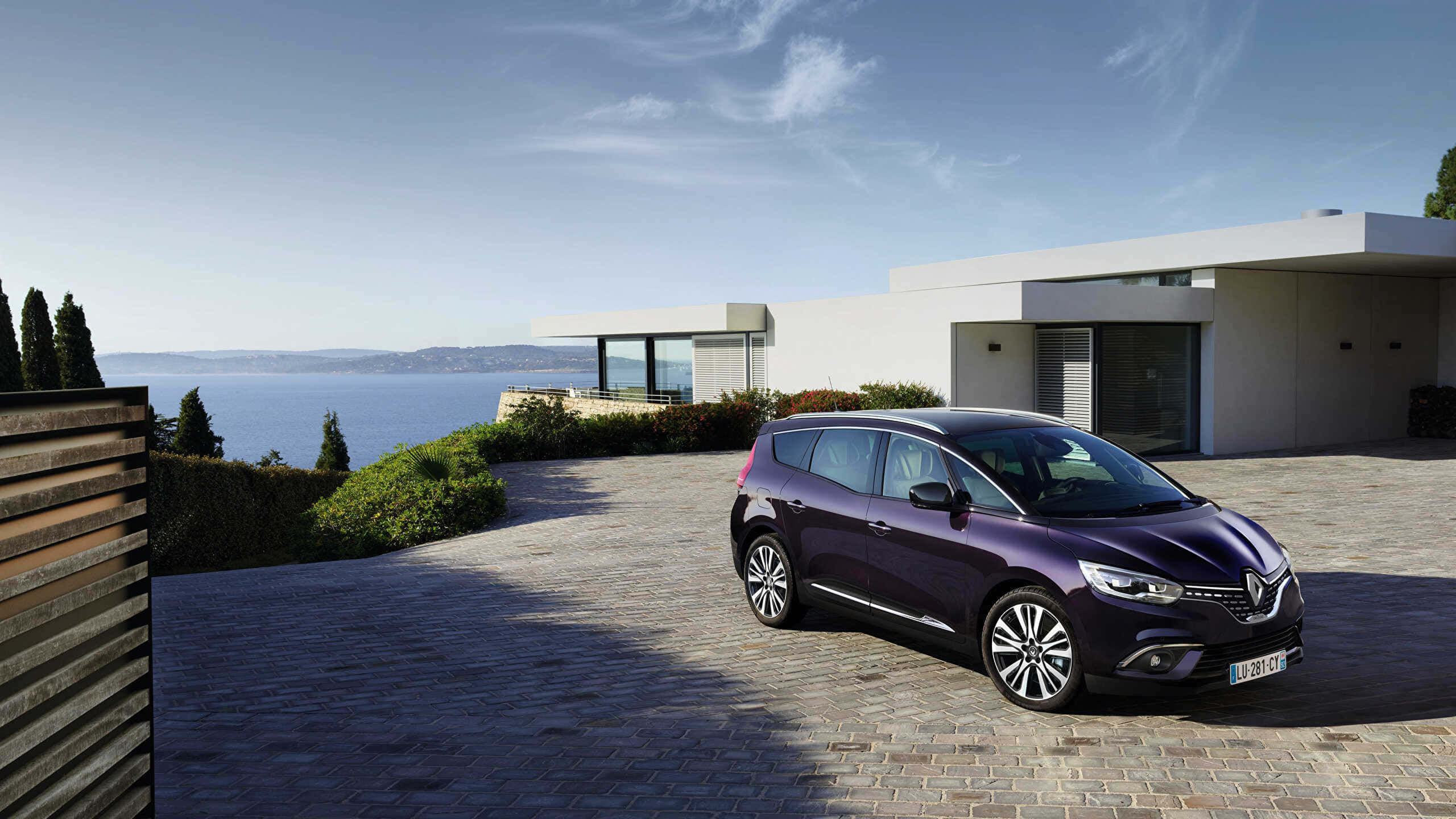 Renault Scenic 桌布 壁紙 — 一架流線型紫色雷諾Scenic停泊在石頭車道上，旁邊是一座現代白色房屋，俯瞰著晴朗天空下的廣闊藍色大海。