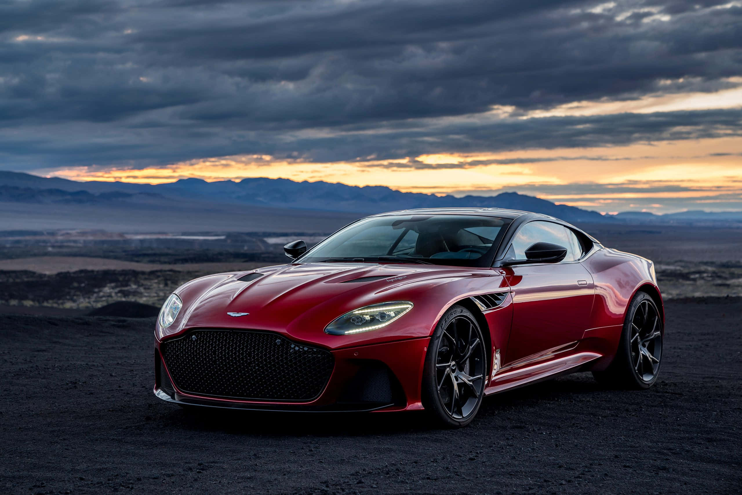 Aston Martin Dbs Superleggera 桌布 壁紙 — 一輛時尚的紅色Aston Martin跑車在戲劇性的暮色天空下閃閃發光，遠處有山巒。
