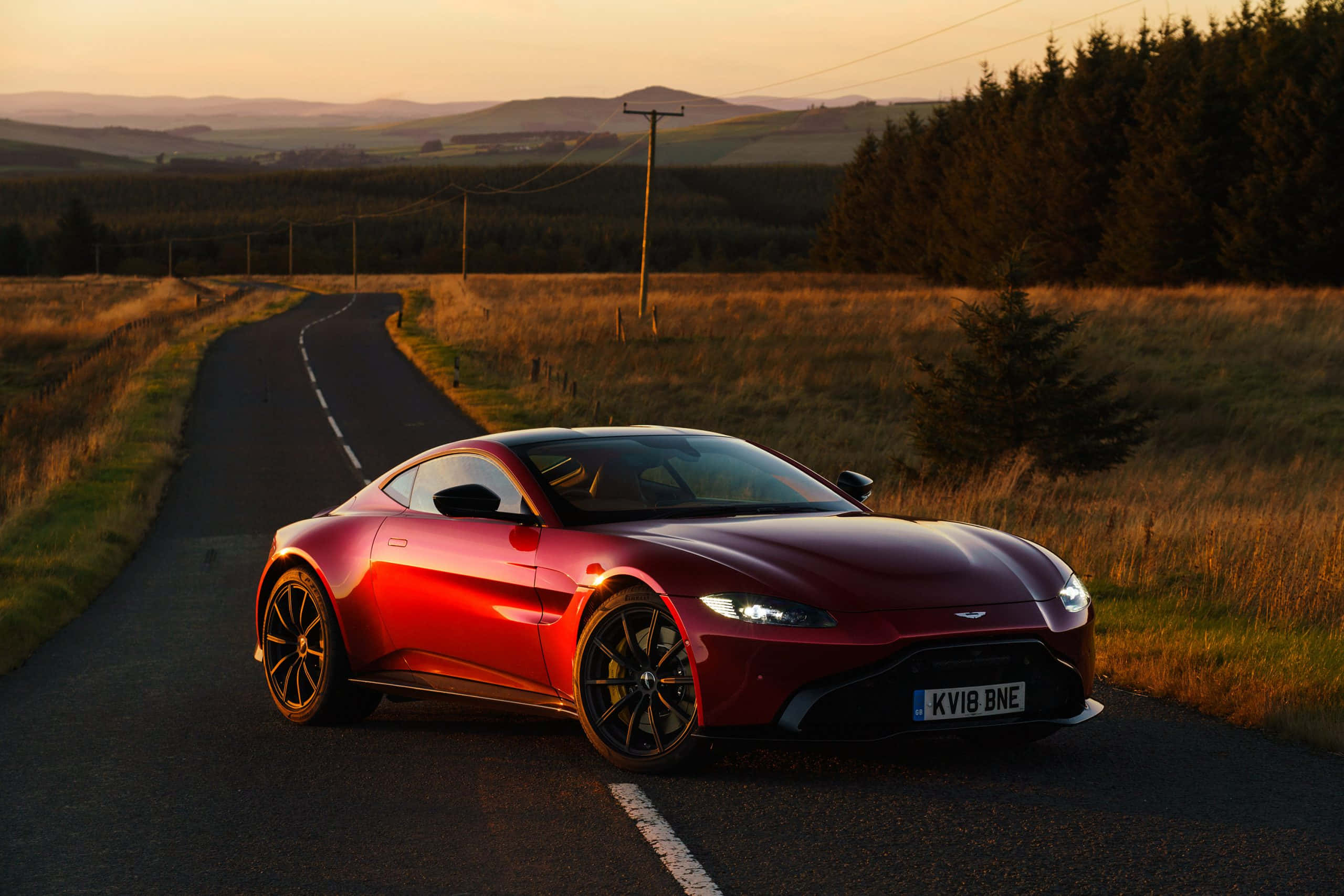 Aston Martin Vantage 桌布 壁紙 — 一輛時尚的紅色跑車在金色日落時分沿著蜿蜒的鄉村道路行駛，背景是起伏的山丘和樹木。