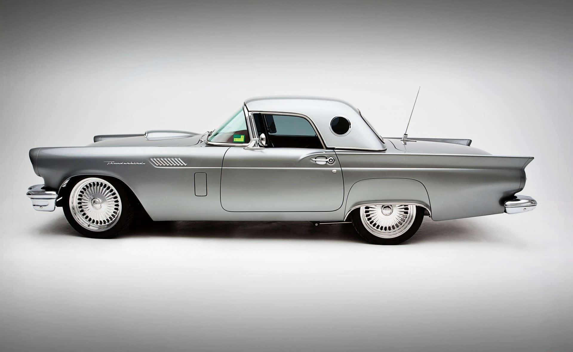 Ford Thunderbird 桌布 壁紙 — 一輛時尚的銀色1950年代福特雷鳥經典車，帶有鍍鉻細節和白牆輪胎，擺放在極簡灰色工作室背景前。