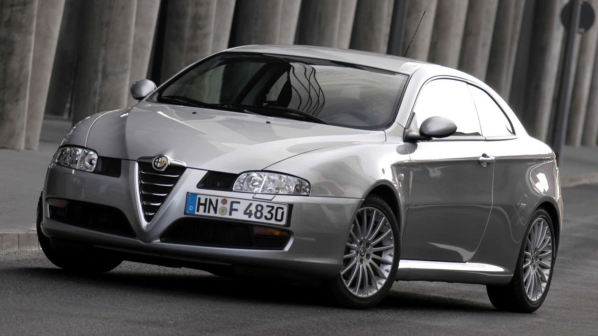 Alfa Romeo Gt 桌布 壁紙 — 一輛時尚的銀色愛快羅密歐雙門跑車停泊在混凝土柱下，配備反光鍍鉻輪圈。