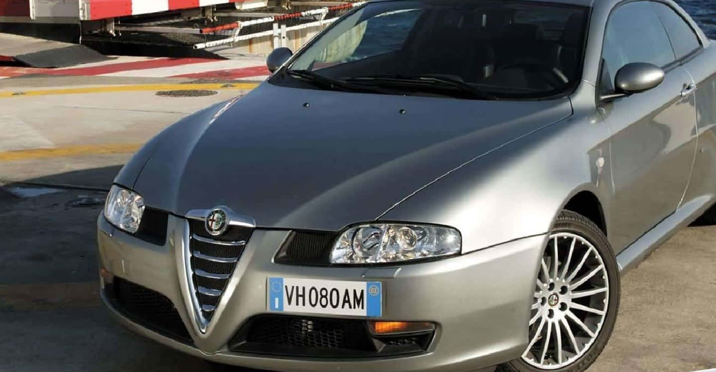 Alfa Romeo Gt 桌布 壁紙 — 一架型格銀色愛快羅密歐跑車雙門轎跑車停泊在混凝土場地上，背景有紅白間條屏障。