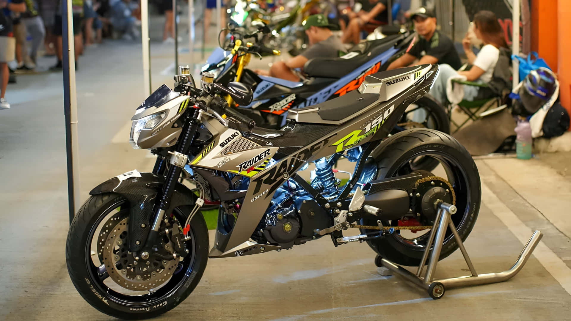  壁紙 — 一輛時尚的銀黑色鈴木 Raider R150 電單車，放在繁忙的室內展覽廳的支架上。