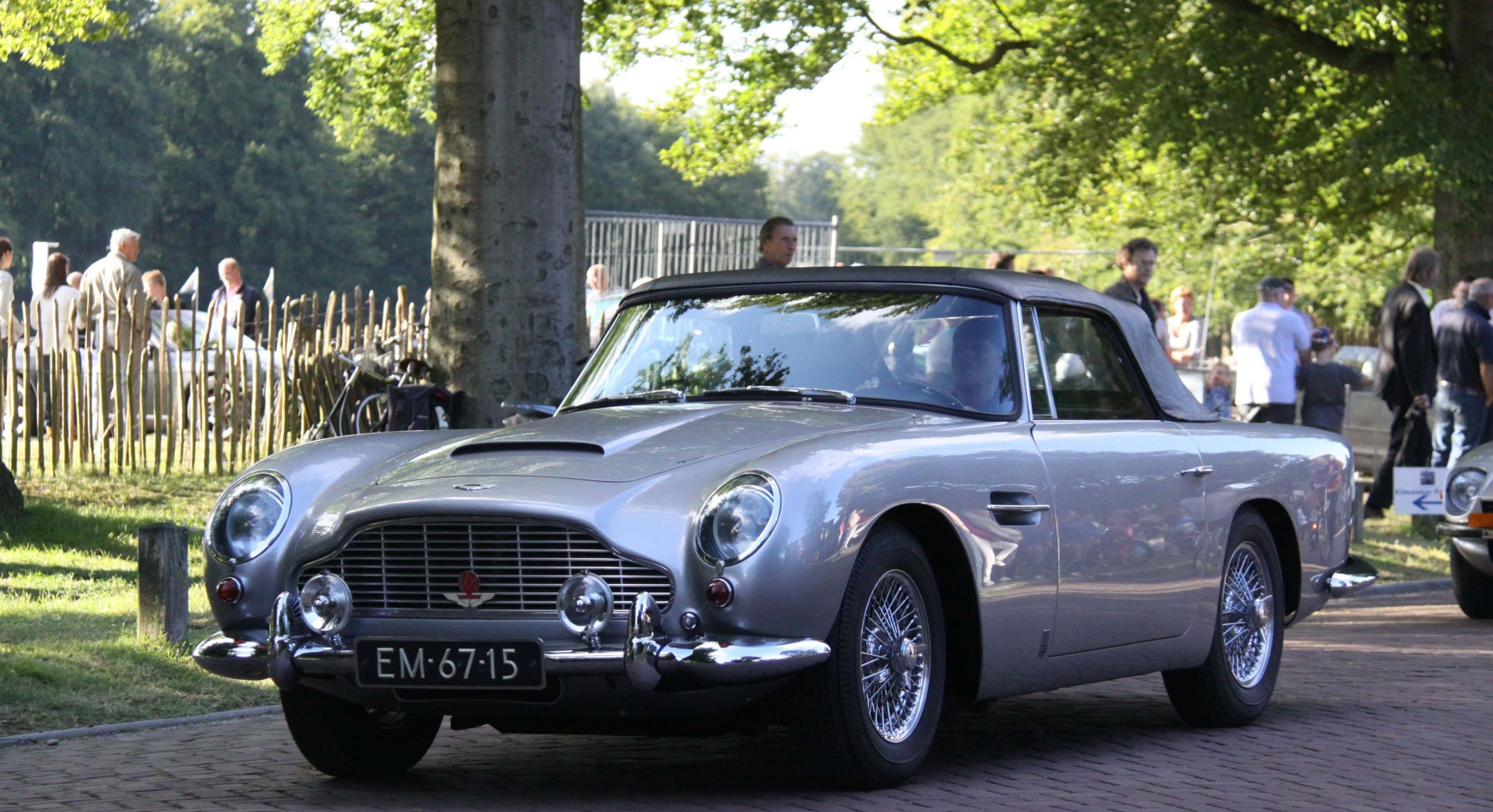 Aston Martin Db5 桌布 壁紙 — 一架時尚的銀色Aston Martin DB5開篷車在明亮陽光下閃閃發光，停泊在磚砌小徑上，周圍是茂密的綠樹和圍觀者。