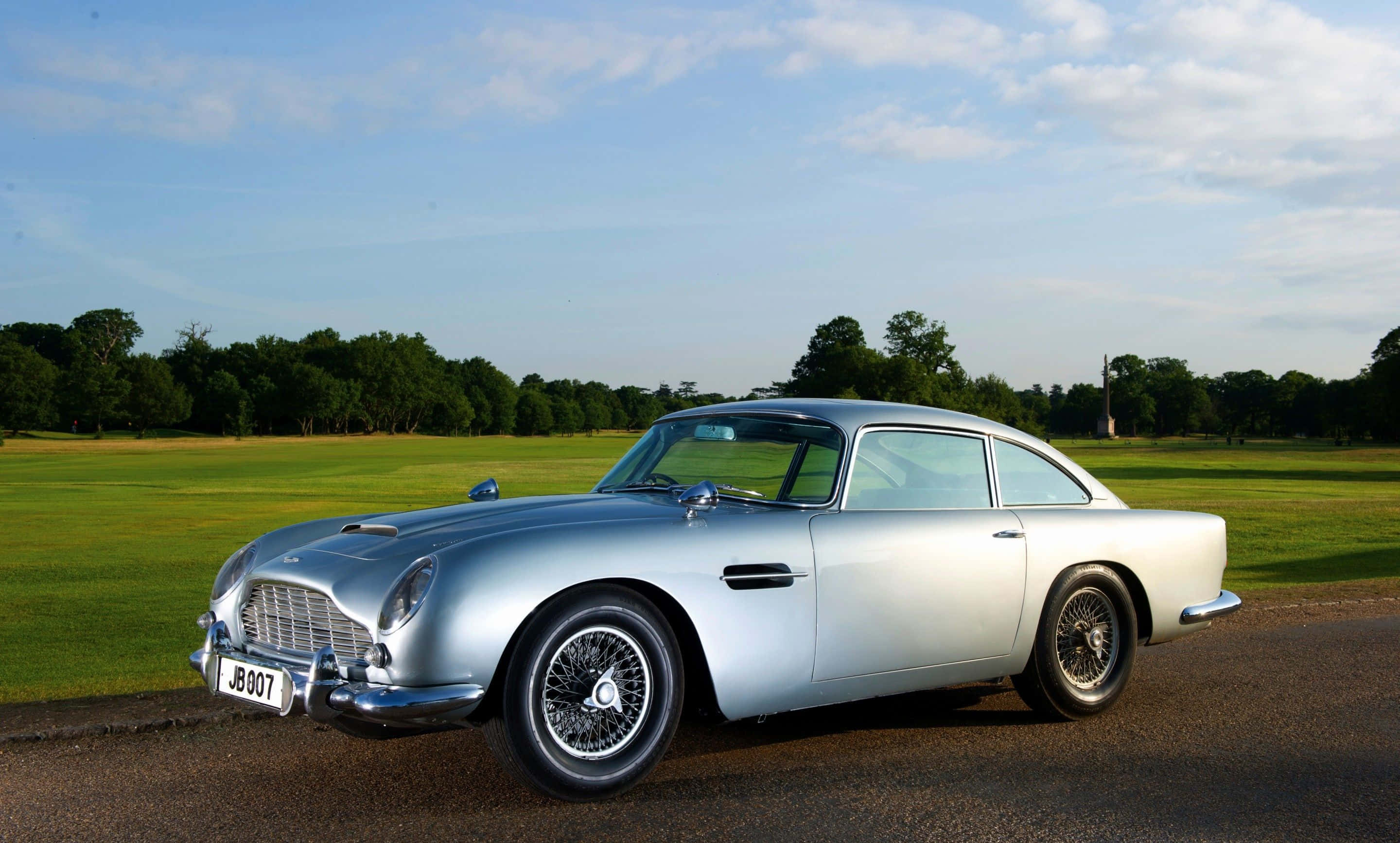 Aston Martin Db5 桌布 壁紙 — 一架流線型的銀色Aston Martin DB5在柔和日光下，於一條鋪砌道路旁廣闊的綠色田野邊閃閃發光。