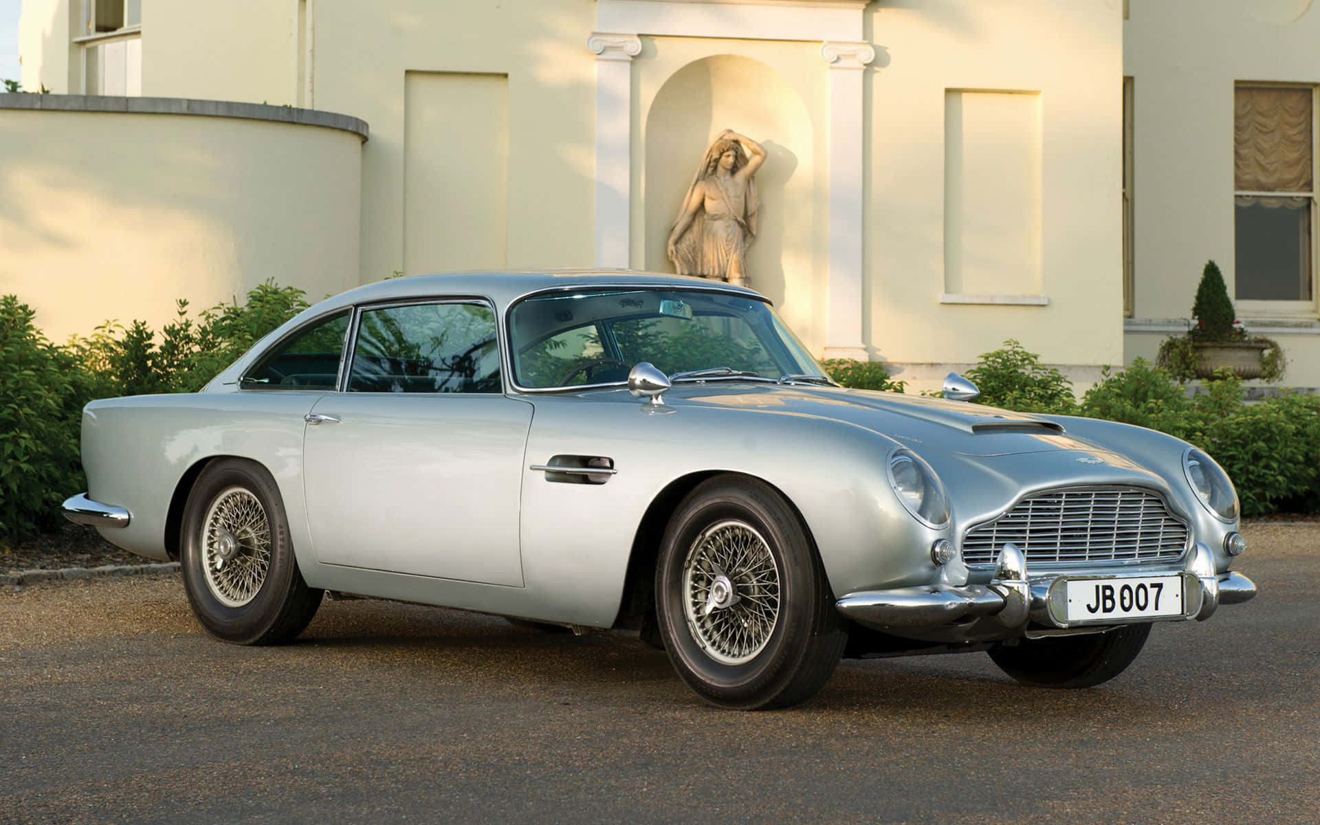Aston Martin Db5 桌布 壁紙 — 一架流線型銀色Aston Martin DB5優雅地停泊在一棟帶有古典雕像的宏偉奶油色大宅前。