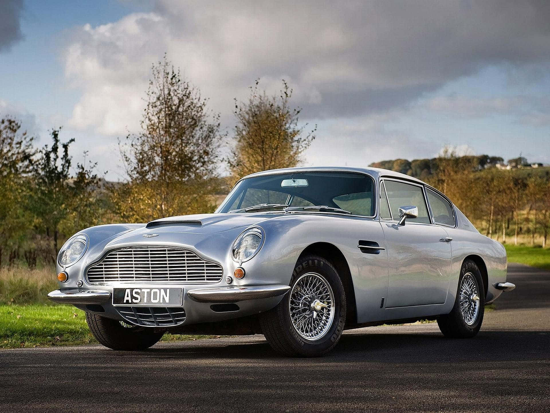 Aston Martin Db5 桌布 壁紙 — 一輛時尚的銀色Aston Martin DB5在多雲的天空下閃閃發光，停在鋪砌的道路上，旁邊是秋天的樹木。