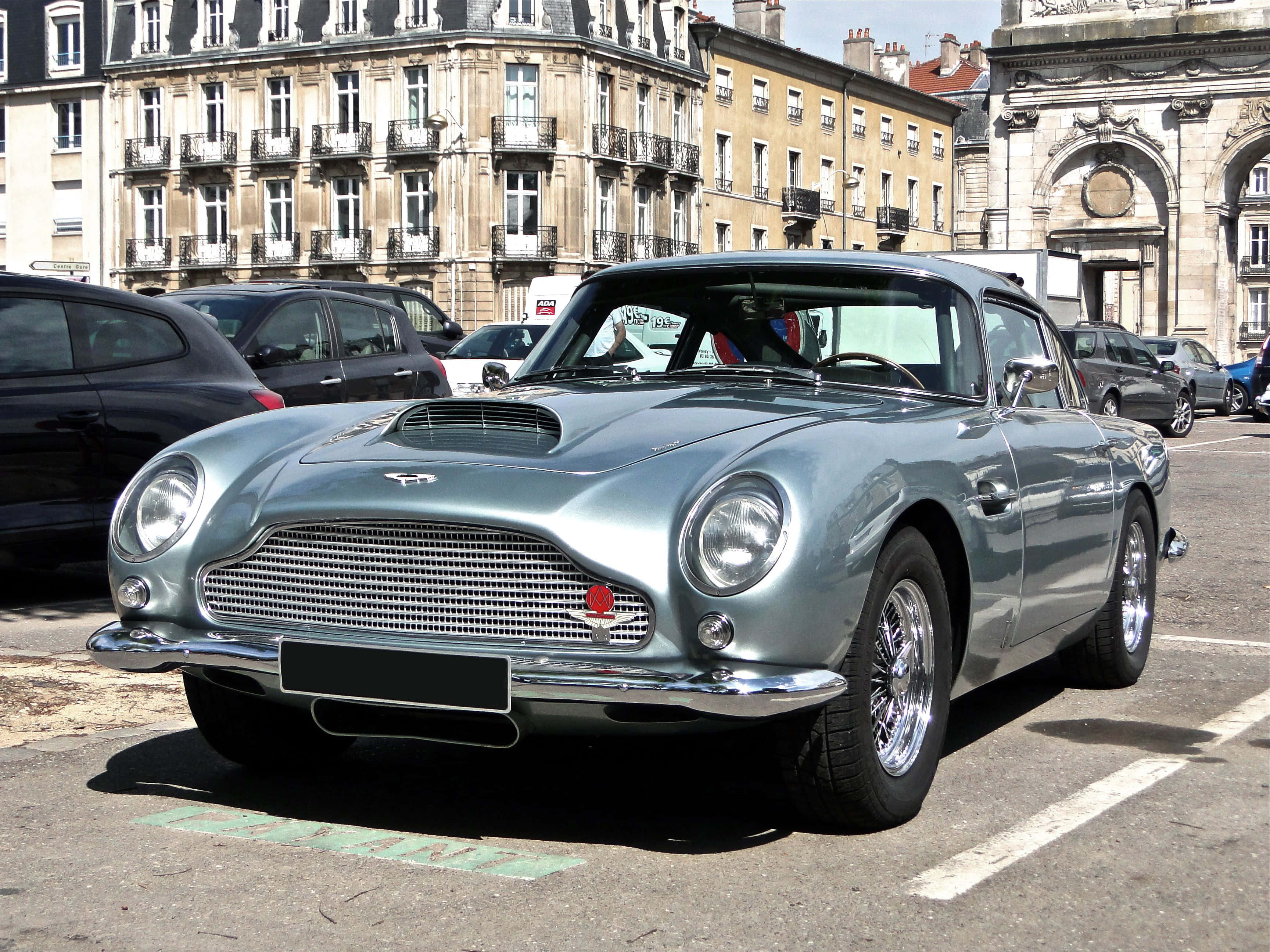 Aston Martin Db5 桌布 壁紙 — 一架時尚的銀色Aston Martin DB6停泊在陽光明媚的歐洲城市廣場，背後是歷史建築。