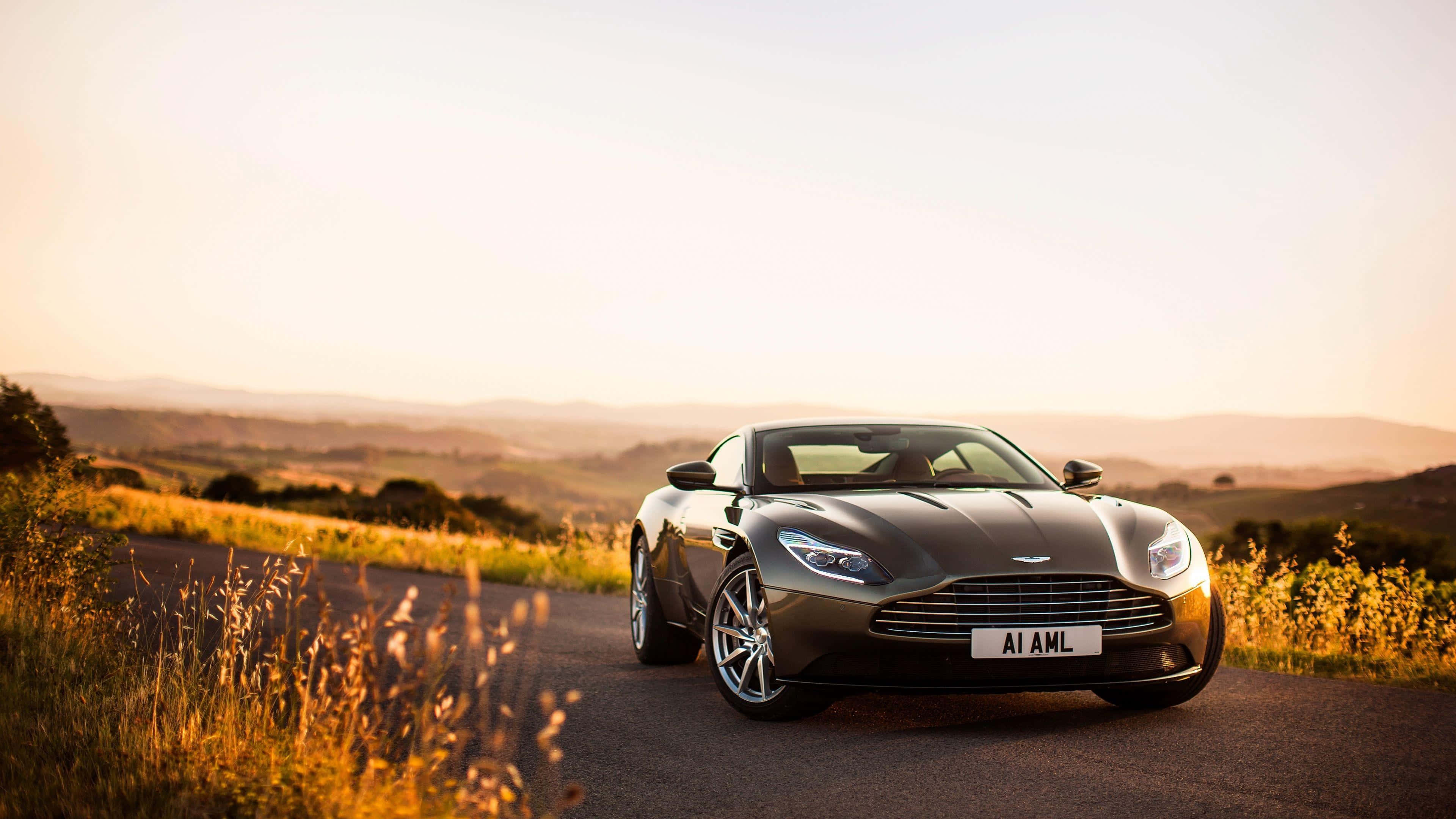Aston Martin Db11 桌布 壁紙 — 一輛時尚的銀色 Aston Martin 跑車停泊在蜿蜒的鄉村道路上，正值黃金時段，背景是起伏的丘陵。