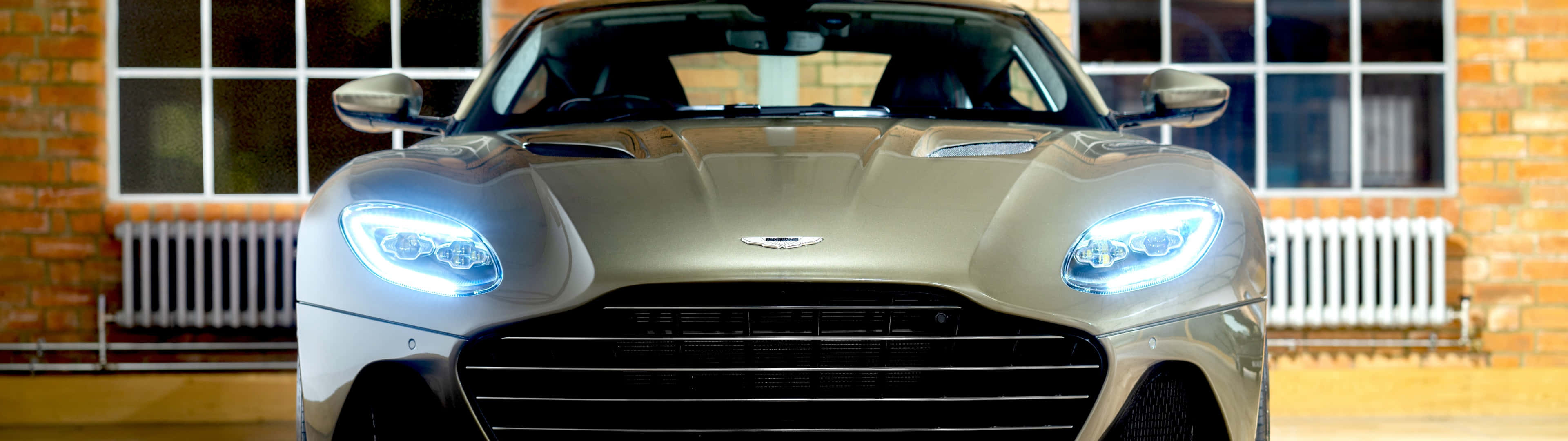 Aston Martin Dbs Superleggera 桌布 壁紙 — 一輛時尚的銀色 Aston Martin 跑車，帶有發光的車頭燈，停在一棟帶有大窗戶的磚砌建築前。