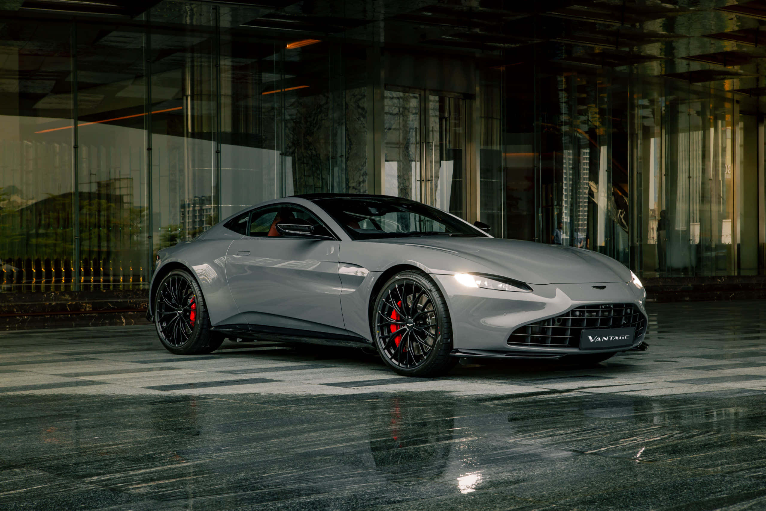 Aston Martin Vantage 桌布 壁紙 — 一輛時尚的銀色Aston Martin Vantage跑車在溫暖的燈光下，在現代玻璃建築旁的潮濕反光路面上閃閃發光。
