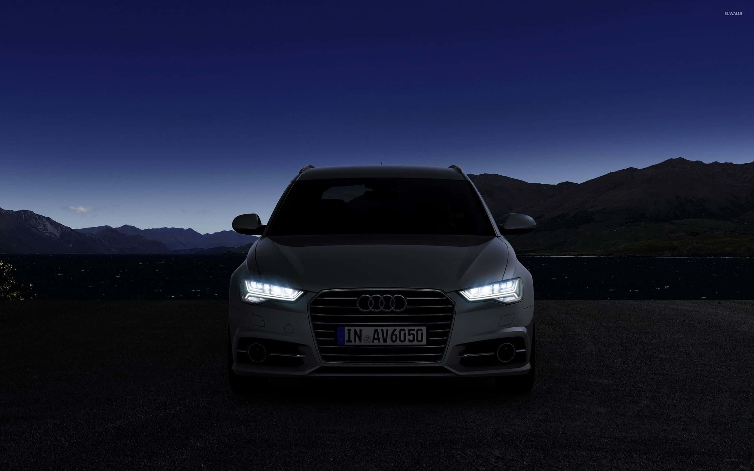 Audi S6 桌布 壁紙 — 一輛時尚的銀色Audi SUV，在暮色中由發光的車頭燈照亮，背景是黑暗的山巒。