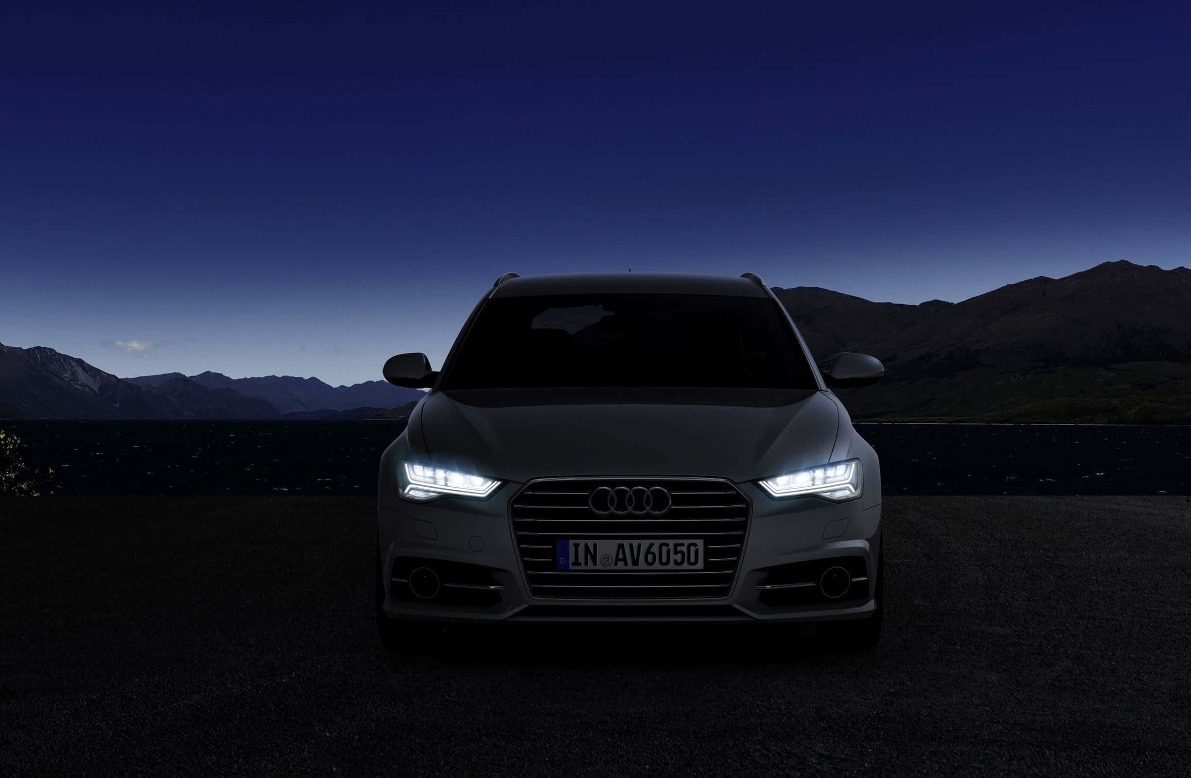 Audi A6 桌布 壁紙 — 一輛時尚的銀色奧迪SUV在暮色天空下，以明亮的車頭燈照亮黑暗的山巒背景。