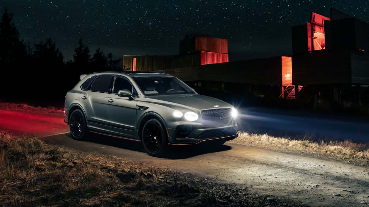 Bentley Bentayga 桌布 壁紙 — 一輛時尚的銀色賓利 Bentayga SUV 頭燈劃破黑夜，在星空下行駛於泥路，經過散發紅光的現代建築。