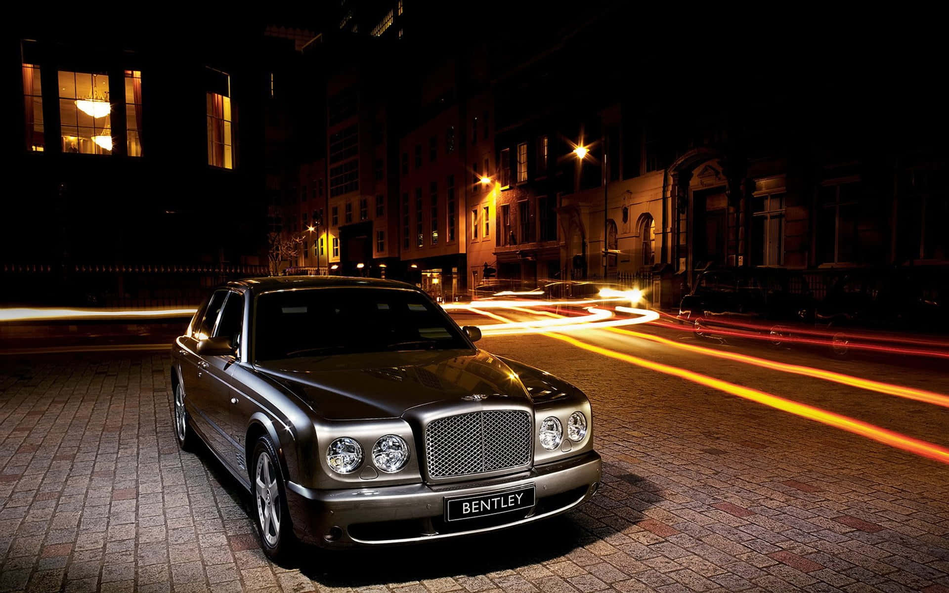 Bentley Arnage 桌布 壁紙 — 一輛時尚的銀色賓利轎車在城市燈光下閃耀，身後是鵝卵石街道上夜間車流的動態模糊痕跡。
