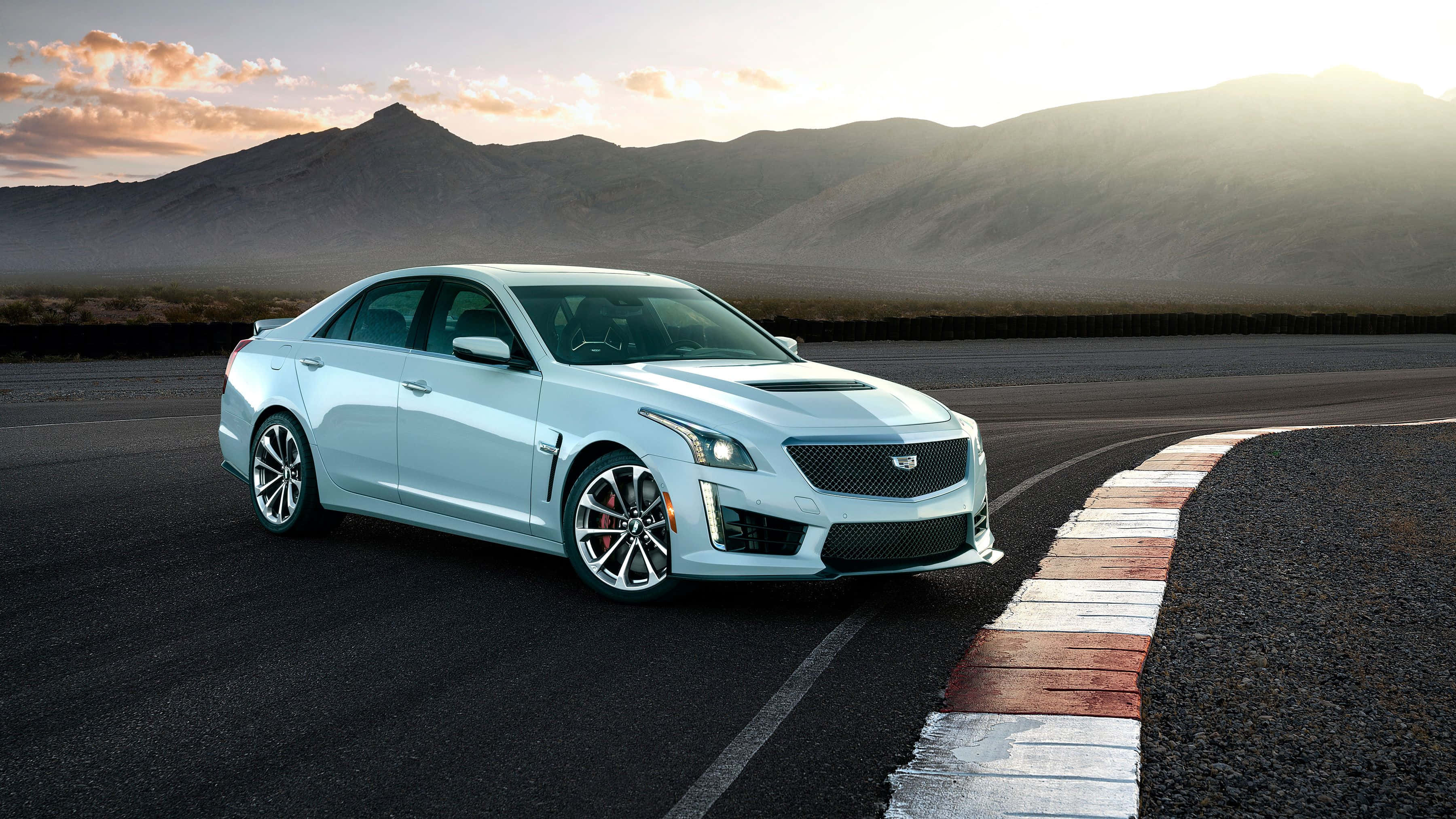 Cadillac Ats 桌布 壁紙 — 一輛時尚的銀色Cadillac CTS-V在日落時分沿著沙漠賽道疾馳，背景是山脈。