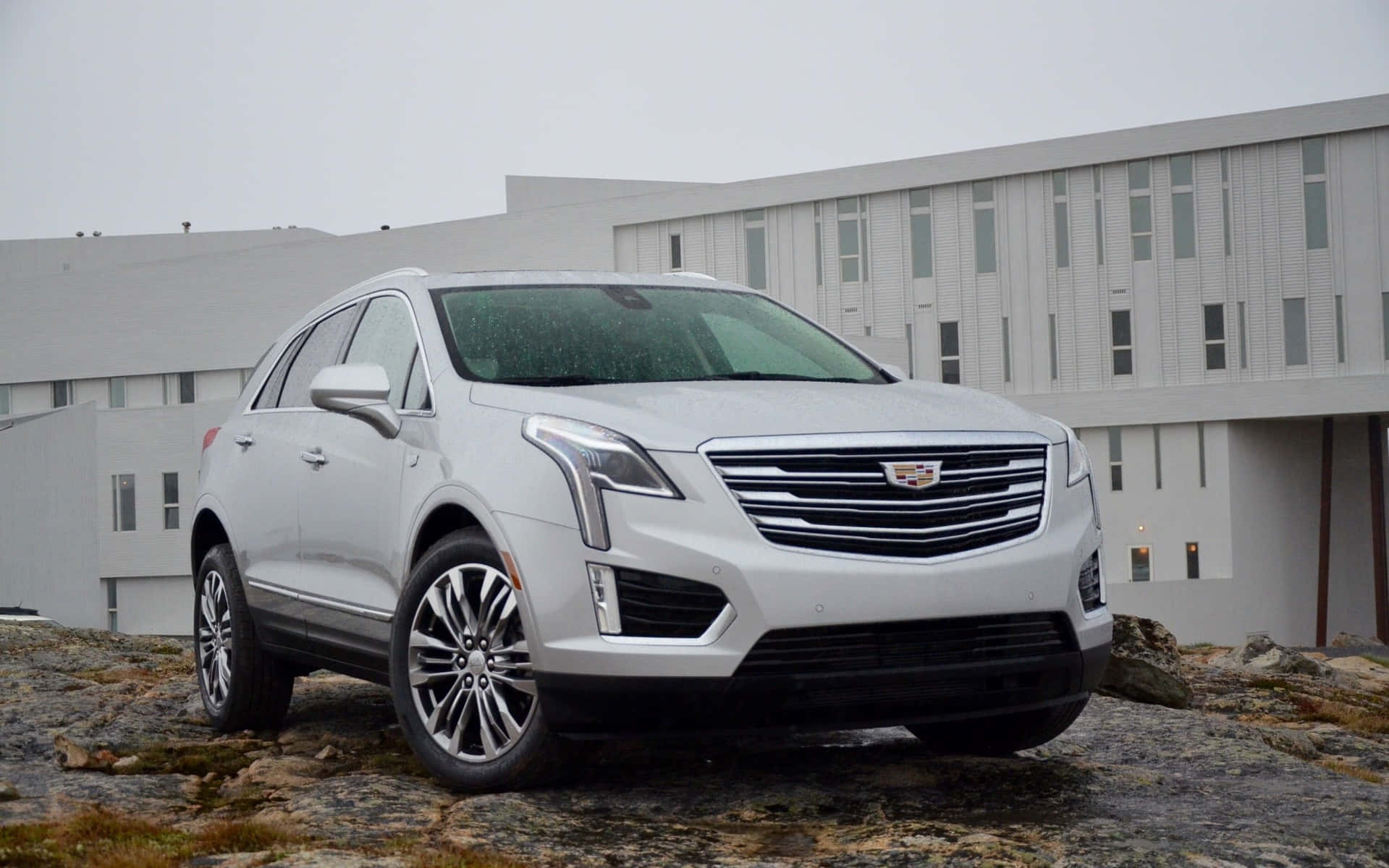 Cadillac Xt5 桌布 壁紙 — 一輛時尚的銀色Cadillac SUV停泊在岩石地形上，前方是一座現代灰色建築，天空多雲。