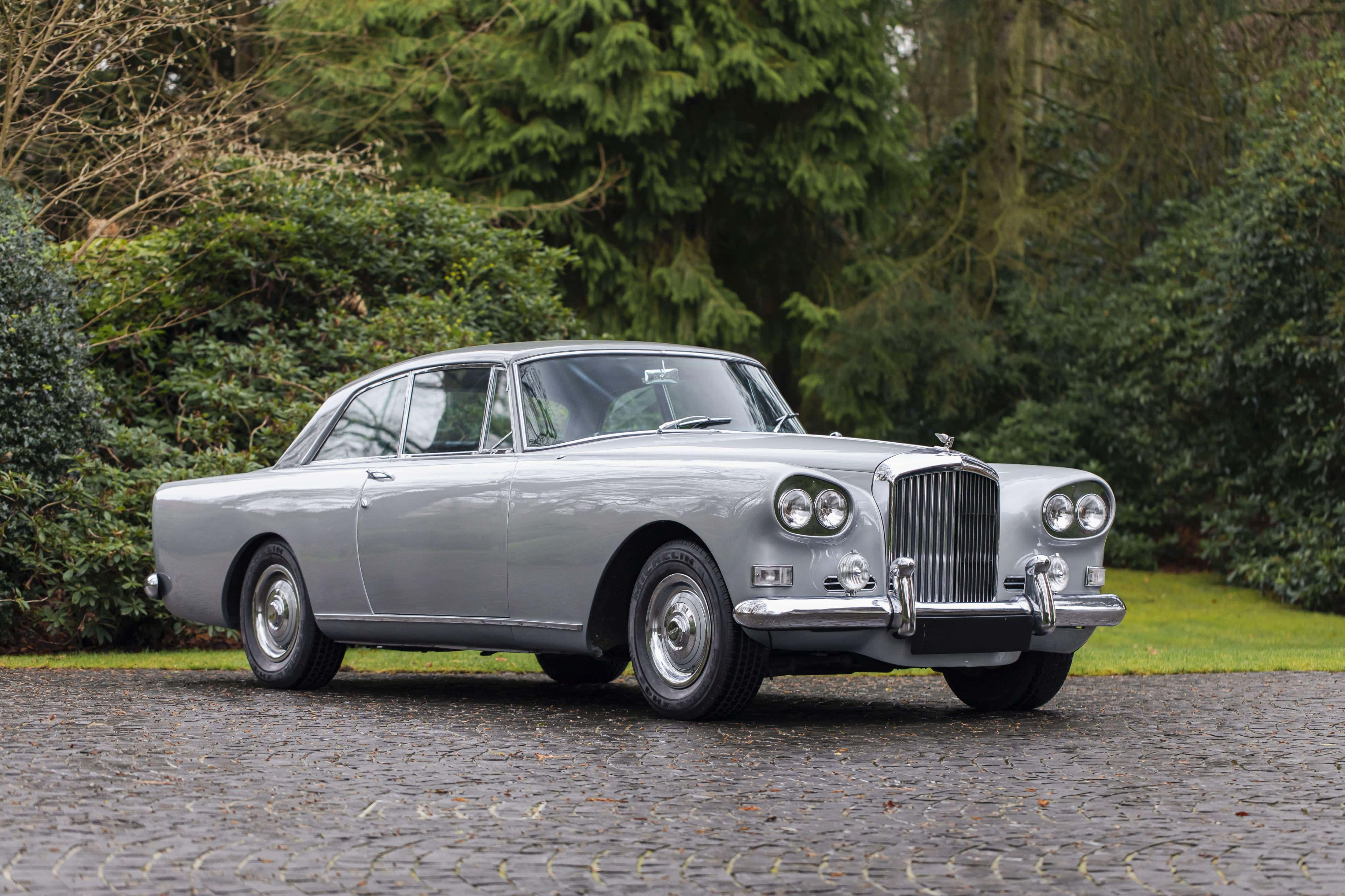 Bentley S3 桌布 壁紙 — 一架時尚的銀色經典賓利轎車停泊在濕漉漉的鵝卵石車道上，周圍環繞著茂密的綠樹。