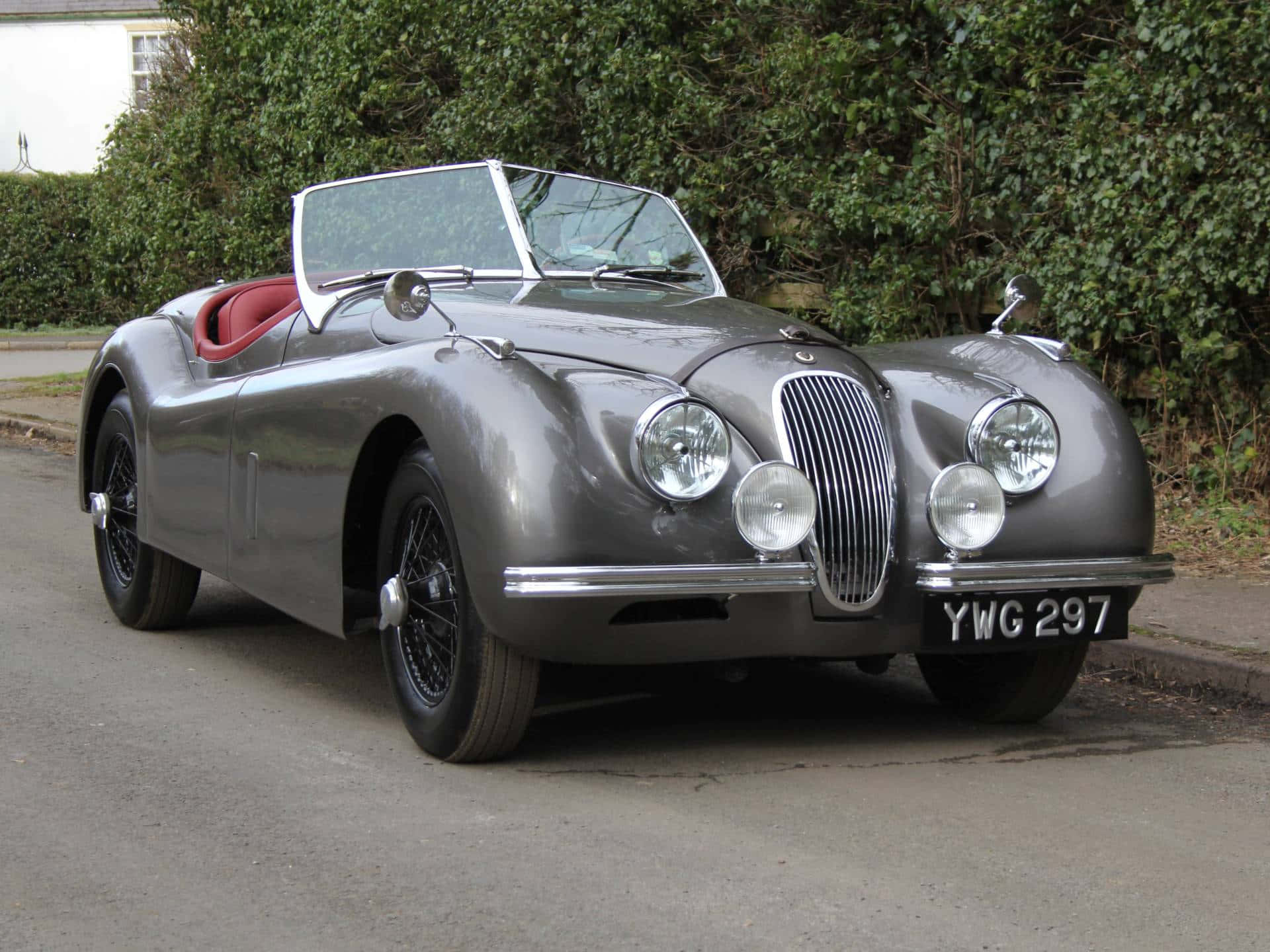  壁紙 — 一架銀色經典Jaguar XK120開篷跑車，配紅色內飾，停泊在寧靜街道旁，旁邊有綠色籬笆。