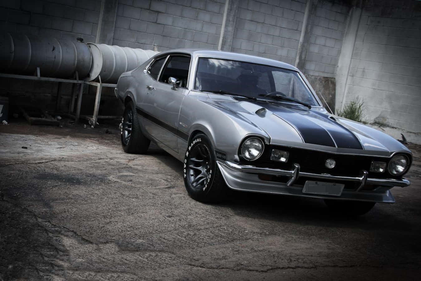 Ford Maverick 桌布 壁紙 — 一架流線型銀色經典肌肉車，帶有黑色賽車條紋，停放在粗獷的工業倉庫中。