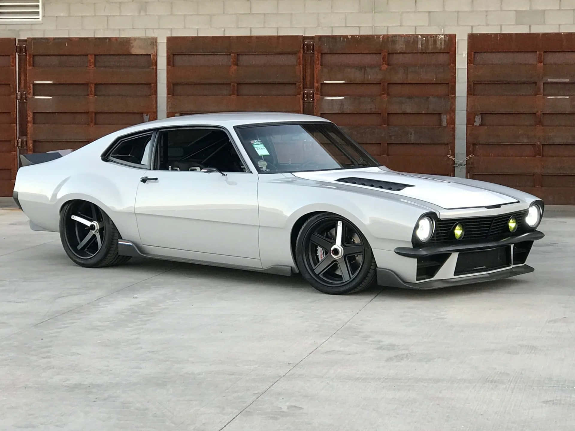 Ford Maverick 桌布 壁紙 — 一輛時尚的銀色經典肌肉車，配備黑色輪圈和發光車頭燈，停在生鏽的木車庫門前。