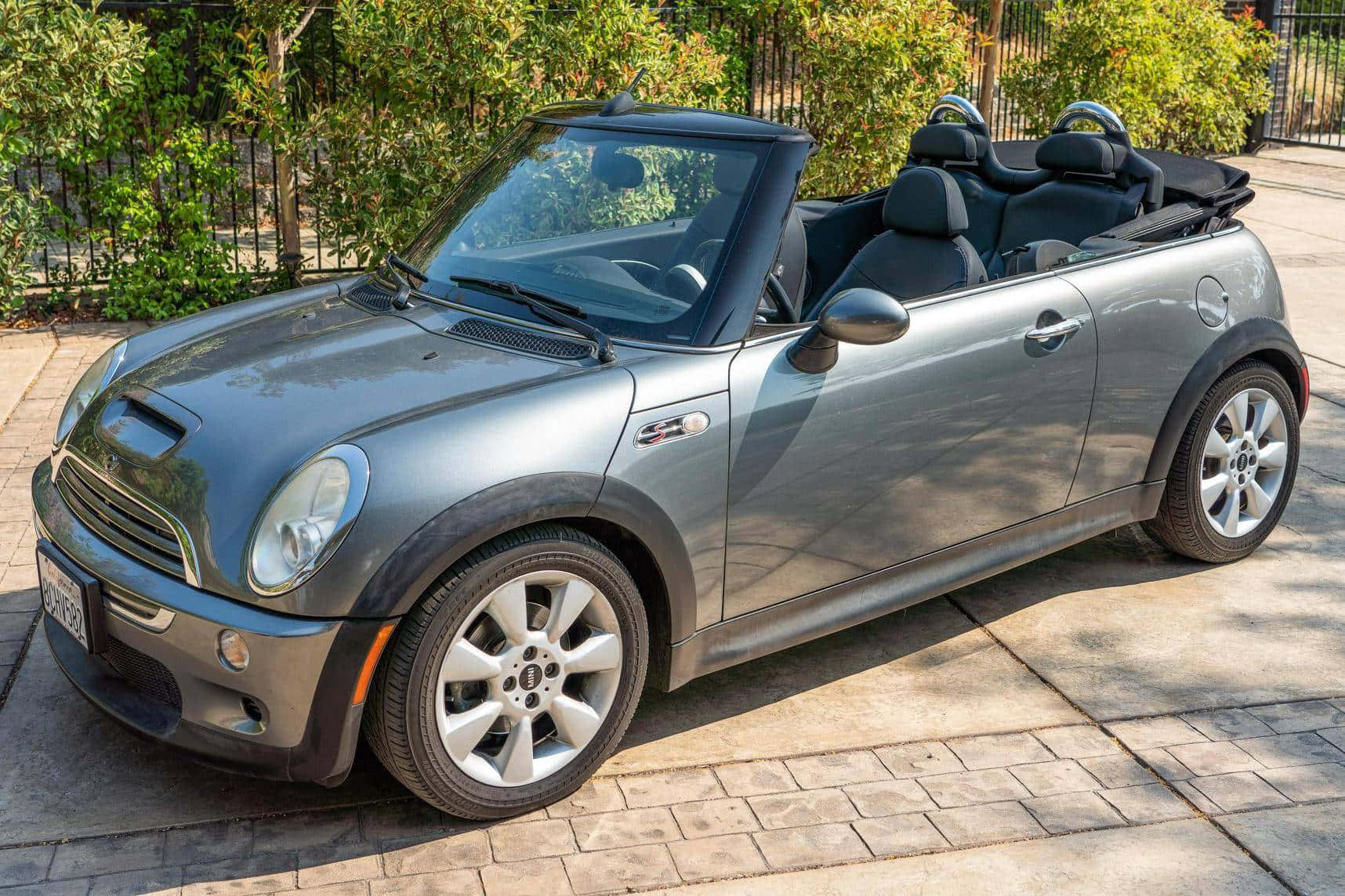 Mini Cooper S Convertible 桌布 壁紙 — 一架時尚的銀色敞篷跑車停泊在陽光普照的石頭車道上，背景是茂密的綠色灌木叢。