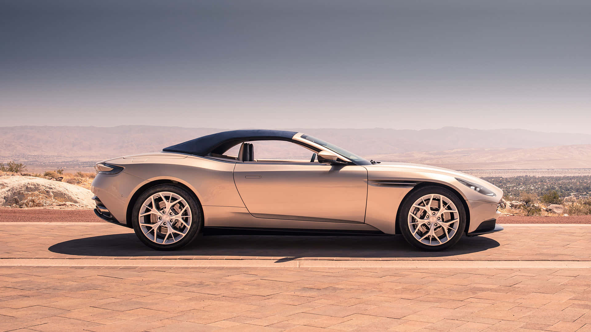 Aston Martin Db11 桌布 壁紙 — 一架時尚的銀色開篷跑車停泊在陽光普照的石頭觀景台上，遠眺朦朧天空下的沙漠山脈。