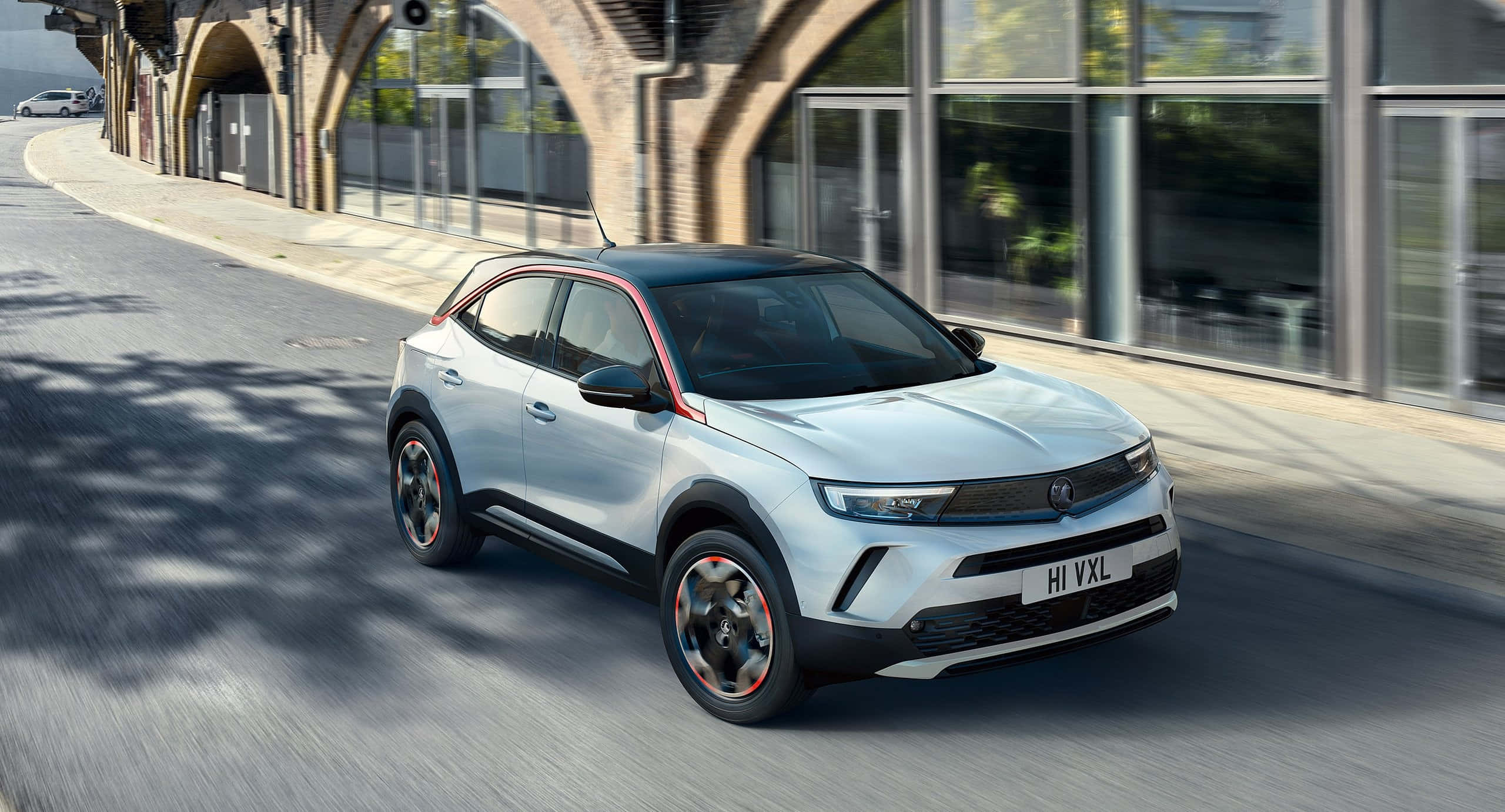 Opel Mokka 桌布 壁紙 — 一輛時尚的銀色電動SUV帶有紅色點綴，在陽光明媚的城市街道上快速行駛於石拱門下。