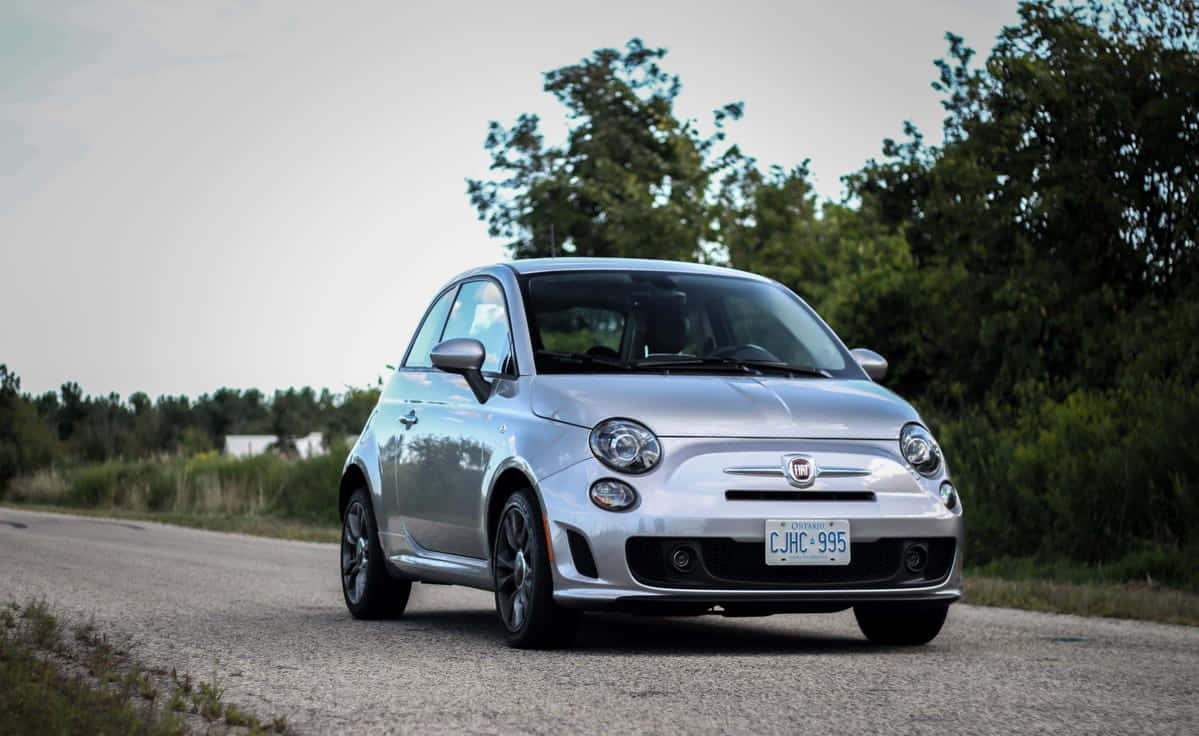 Fiat 500 桌布 壁紙 — 一輛時尚的銀色Fiat 500在陰天下行駛在一條兩旁綠樹成蔭的寧靜鄉村道路上。