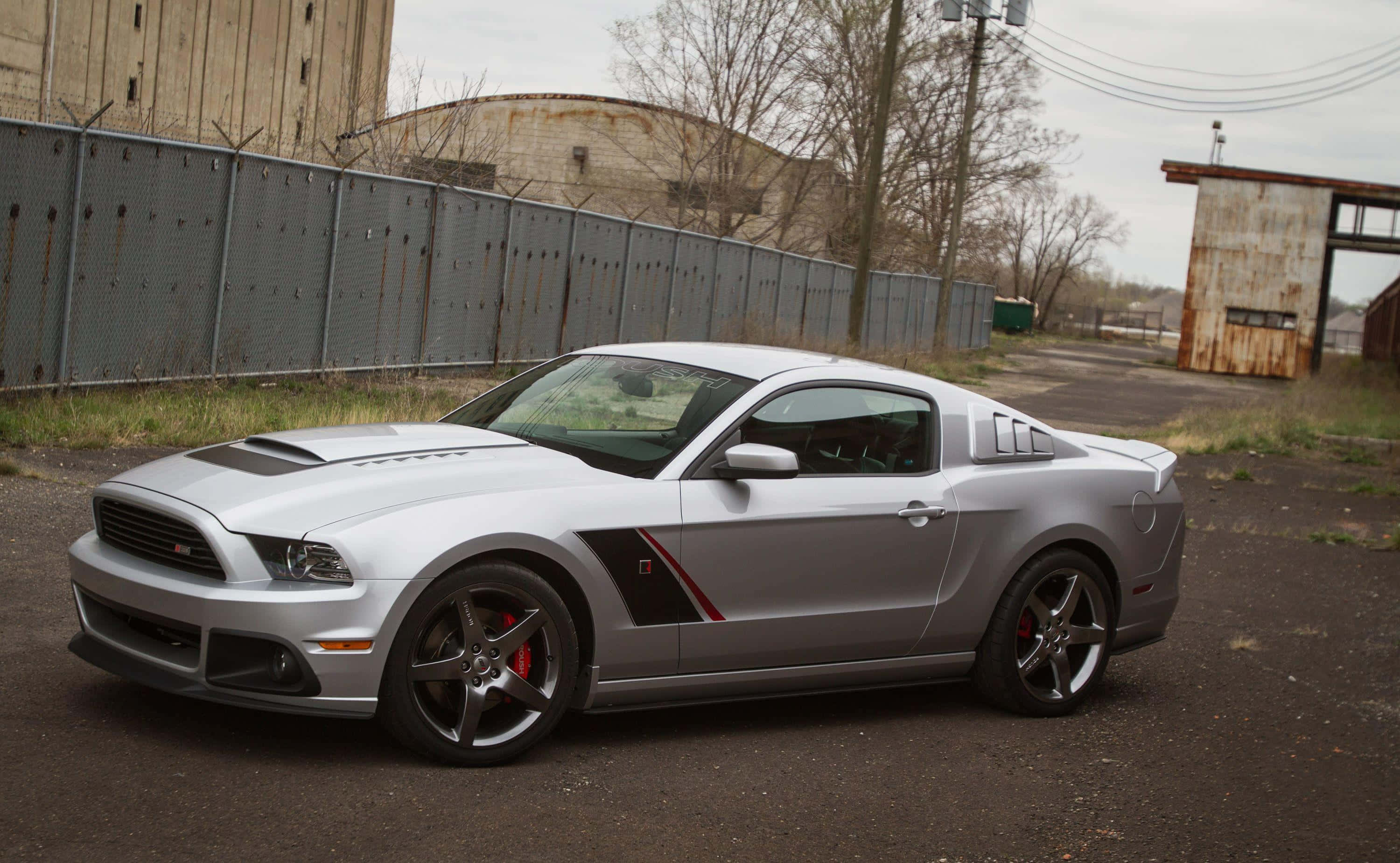 Ford Mustang Roush 桌布 壁紙 — 一架時尚的銀色福特野馬Shelby GT500停泊在工業用地，周圍有生鏽的圍欄和倉庫，天空多雲。