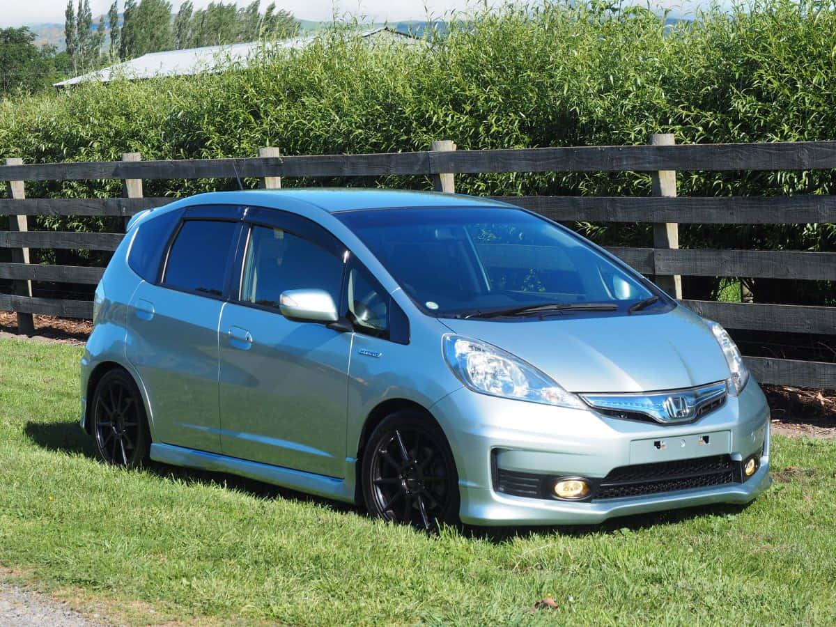 Honda Fit 桌布 壁紙 — 一架時尚的銀色本田Fit停泊在草地旁，旁邊有木柵欄，背景是茂密的綠色樹籬。