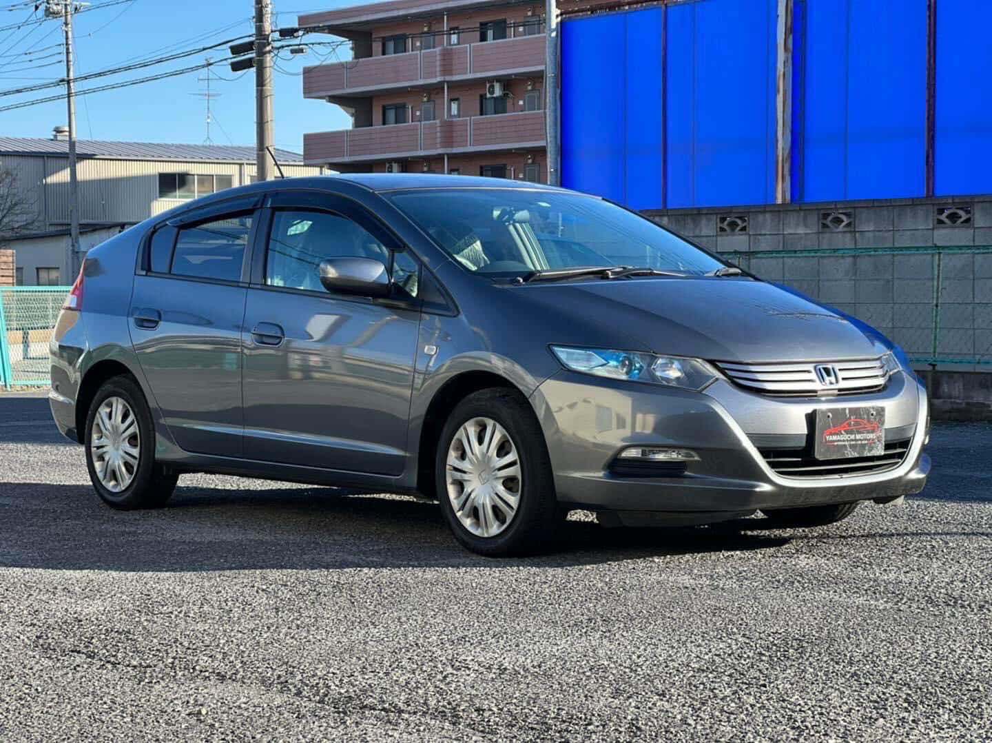Honda Insight 桌布 壁紙 — 一輛時尚的銀色本田Insight混合動力車停在碎石上，背景有藍色牆壁和公寓大樓，在晴朗的日光下。