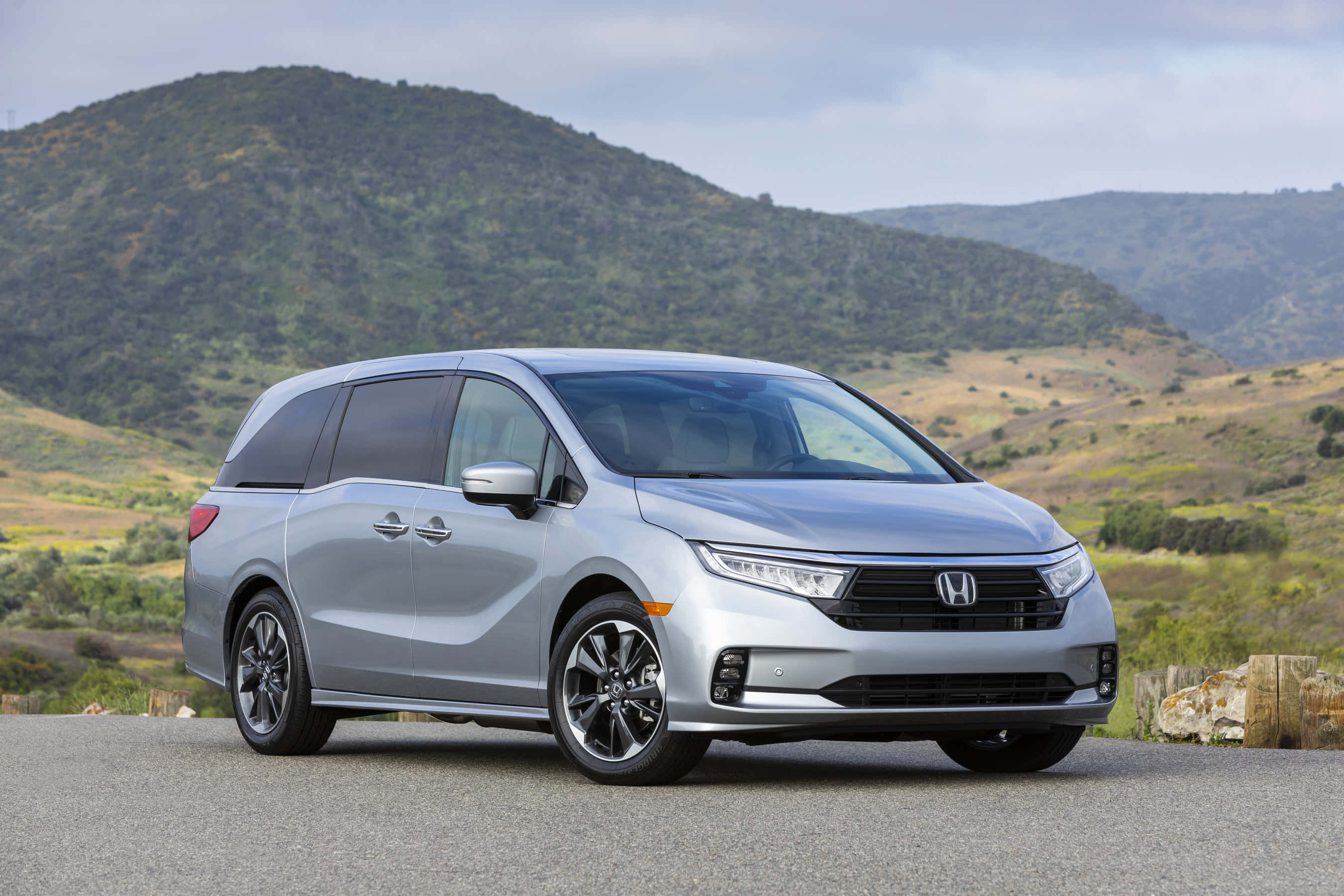 Honda Odyssey 桌布 壁紙 — 一架流線型銀色本田小型貨車停泊在鋪砌道路上，背景是起伏的綠色山丘和多雲的天空。