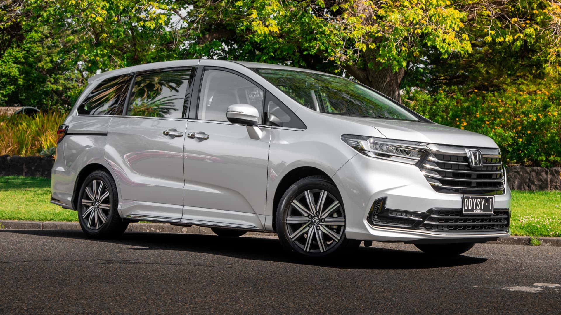 Honda Odyssey 桌布 壁紙 — 一架流線型銀色本田Odyssey小型貨車停泊在鋪砌道路上，背景有茂密的綠樹和黃花。