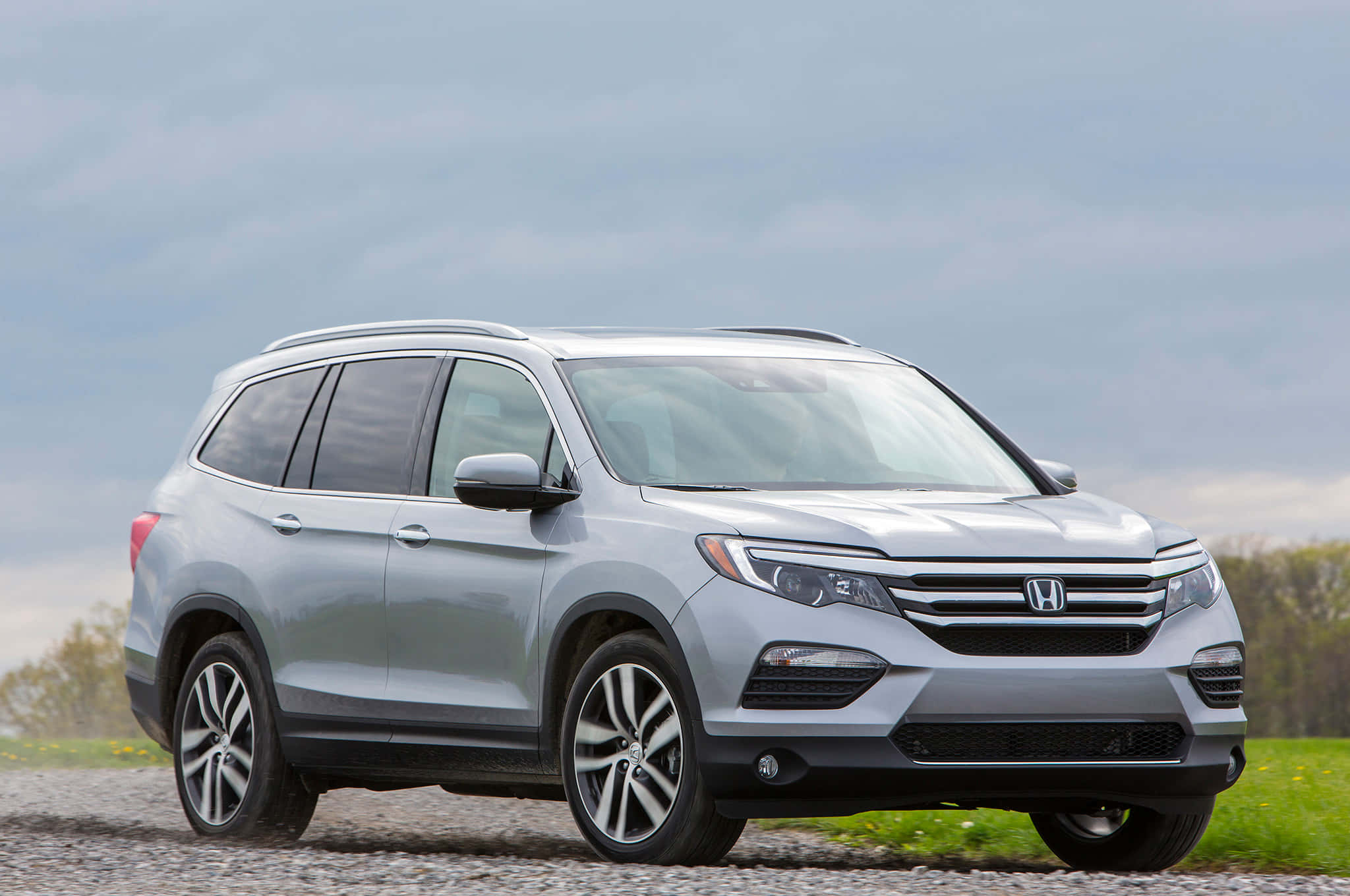 Honda Pilot 桌布 壁紙 — 一架時尚的銀色本田Pilot SUV停泊在碎石路上，背景有綠草和陰天。