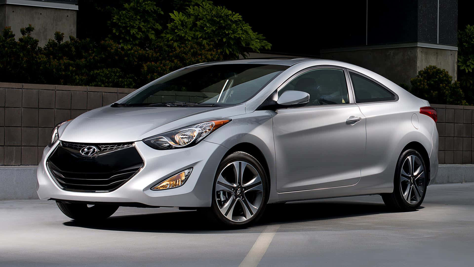 Hyundai Elantra 桌布 壁紙 — 一架時尚的銀色現代轎跑車停在混凝土表面上，背景有深色樹葉和混凝土牆。