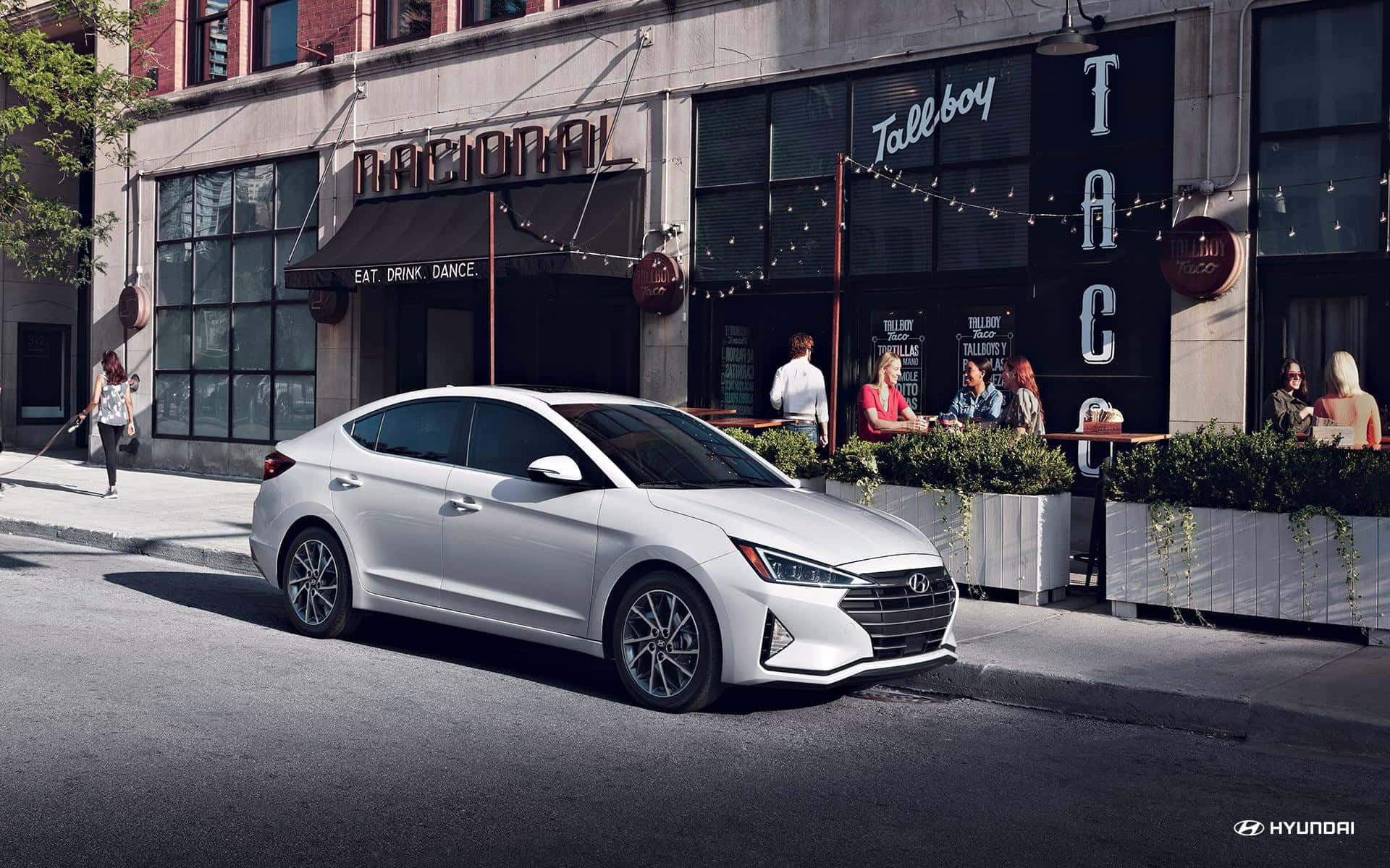 Hyundai Elantra 桌布 壁紙 — 一架時尚的銀色現代Elantra停泊在陽光明媚的城市街道上，旁邊是一間設有戶外座位的熱鬧墨西哥卷餅餐廳。