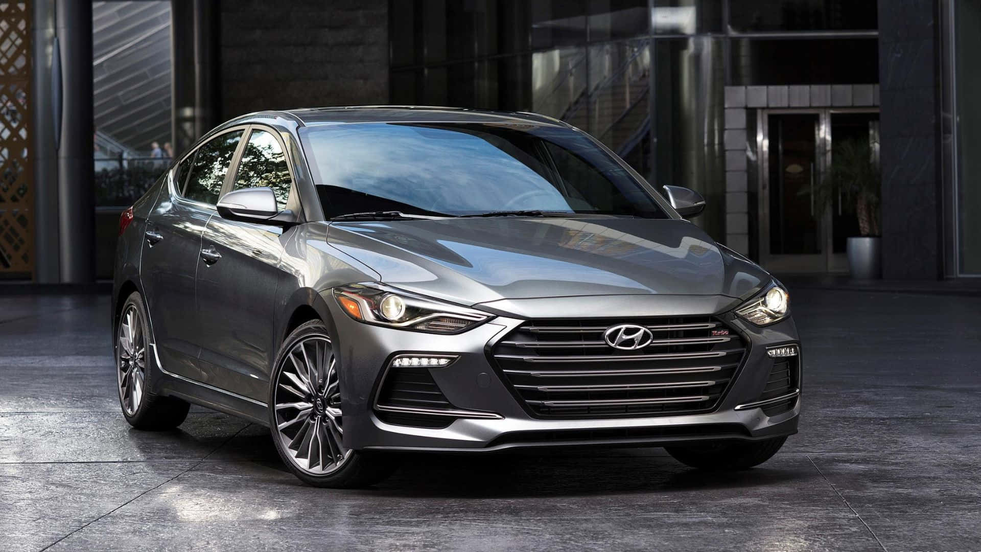 Hyundai Elantra 桌布 壁紙 — 一輛時尚的銀色現代伊蘭特轎車在明亮的日光下閃閃發光，停在現代玻璃和鋼結構建築前。