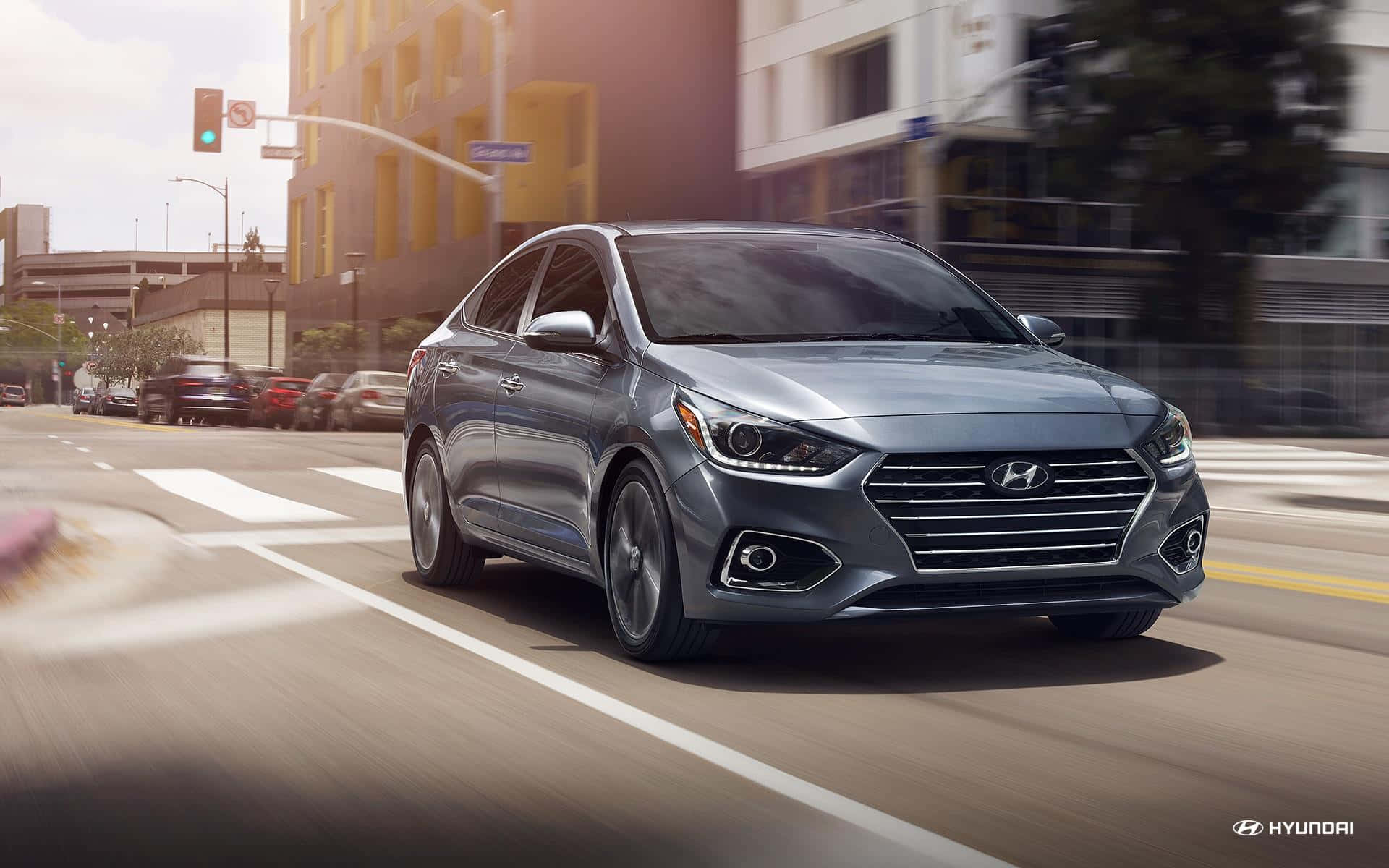 Hyundai Accent 桌布 壁紙 — 一輛時尚的銀色現代房車在陽光普照的城市街道上飛馳，背景建築物和道路呈現動態模糊效果。