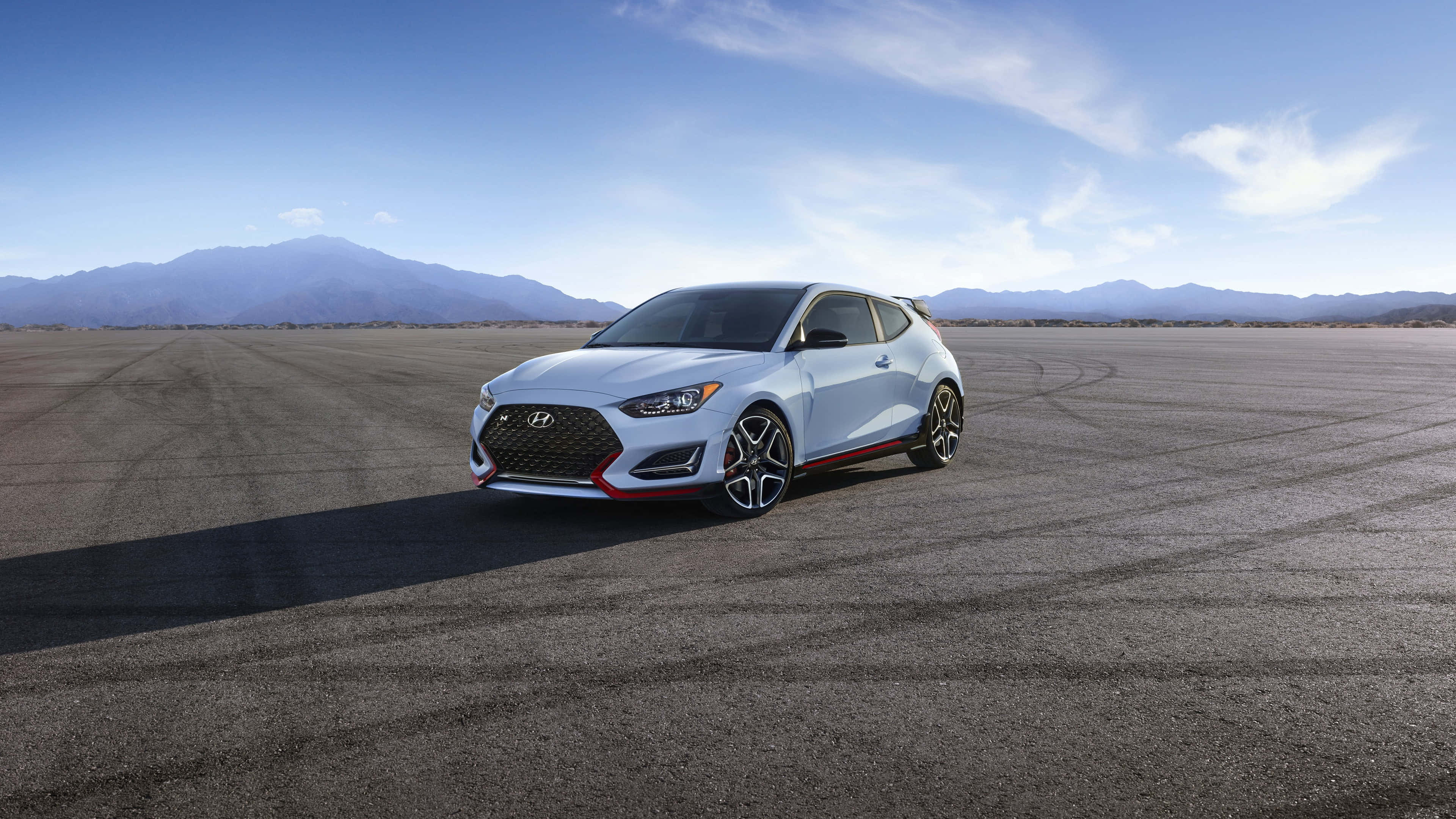 Hyundai Veloster 桌布 壁紙 — 一輛時尚的銀色現代Veloster N跑車，帶有紅色點綴，停在廣闊的瀝青賽道上，背景是晴朗的藍天和遠處的山脈。