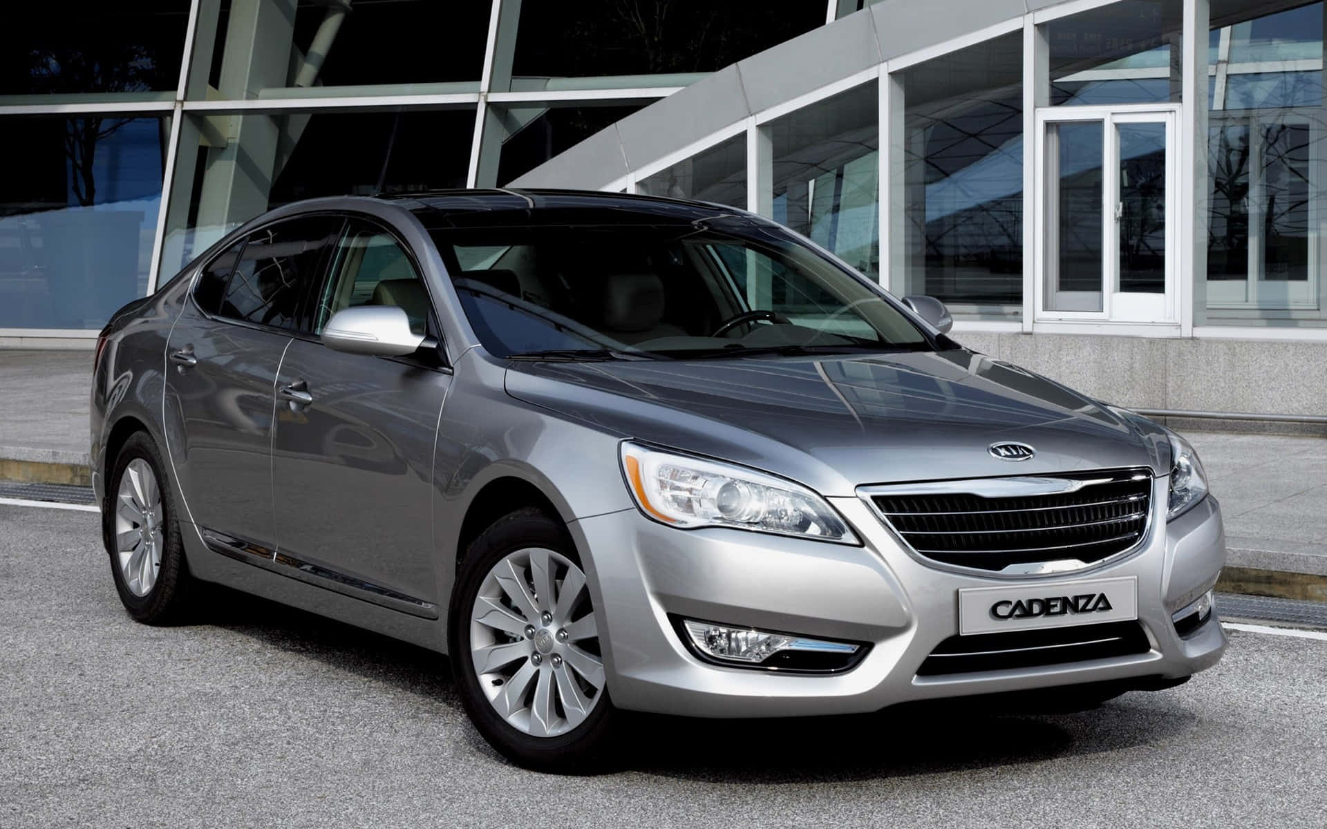 Kia Cadenza 桌布 壁紙 — 一輛時尚的銀色Kia Cadenza轎車在晴朗的日子停在一棟現代玻璃建築前。