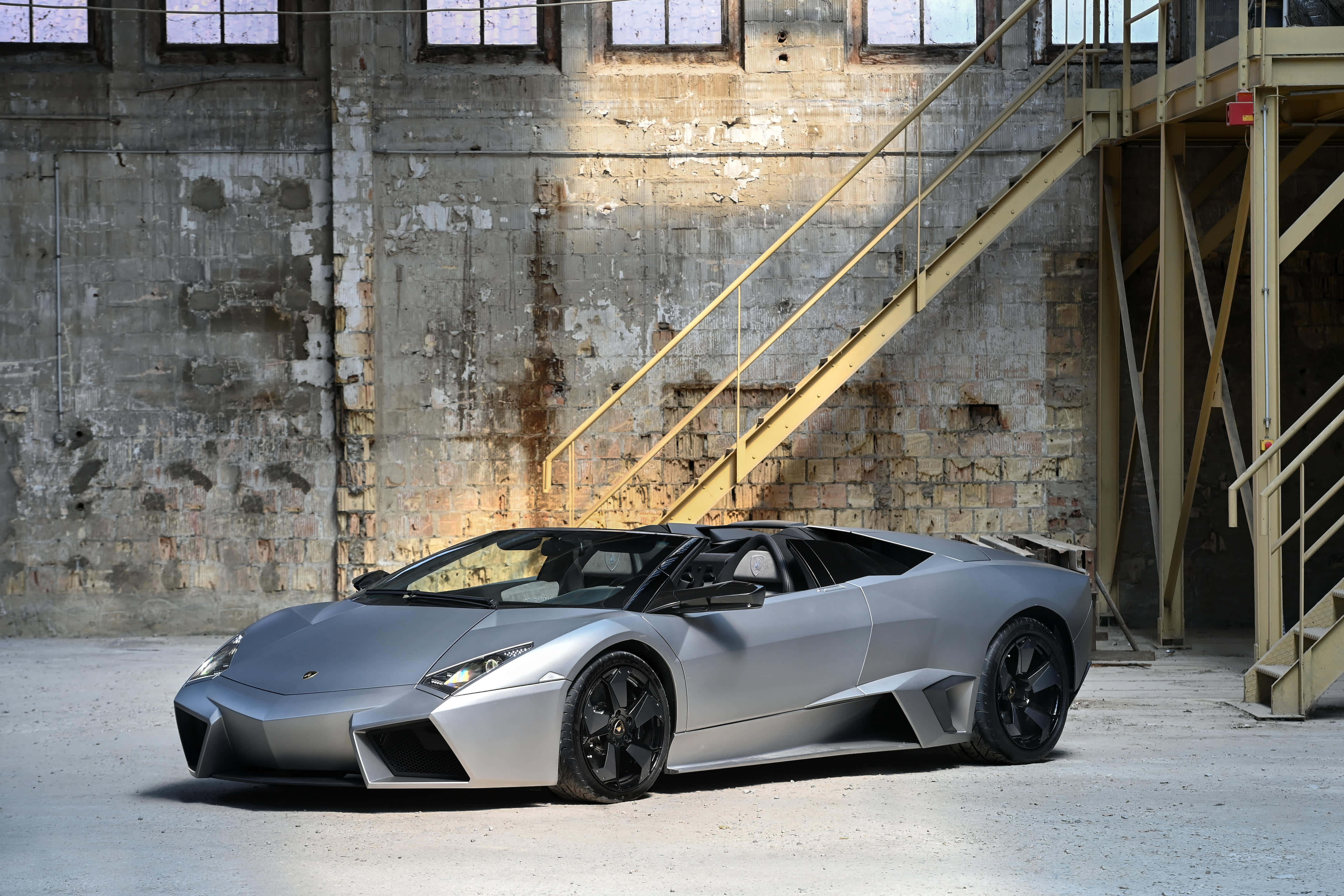 Lamborghini Reventón 桌布 壁紙 — 一架時尚的銀色林寶堅尼 Reventon Roadster 停泊在一個風化的工業倉庫內，倉庫有外露的混凝土牆和黃色金屬樓梯。