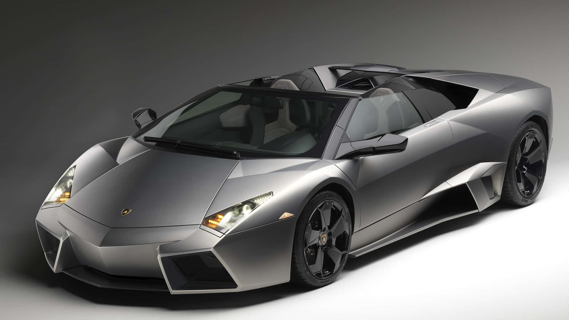 Lamborghini Reventón 桌布 壁紙 — 一部時尚的銀色林寶堅尼 Reventon Roadster，帶有黑色飾邊和發光的車頭燈，在簡約的灰色工作室背景下擺拍。