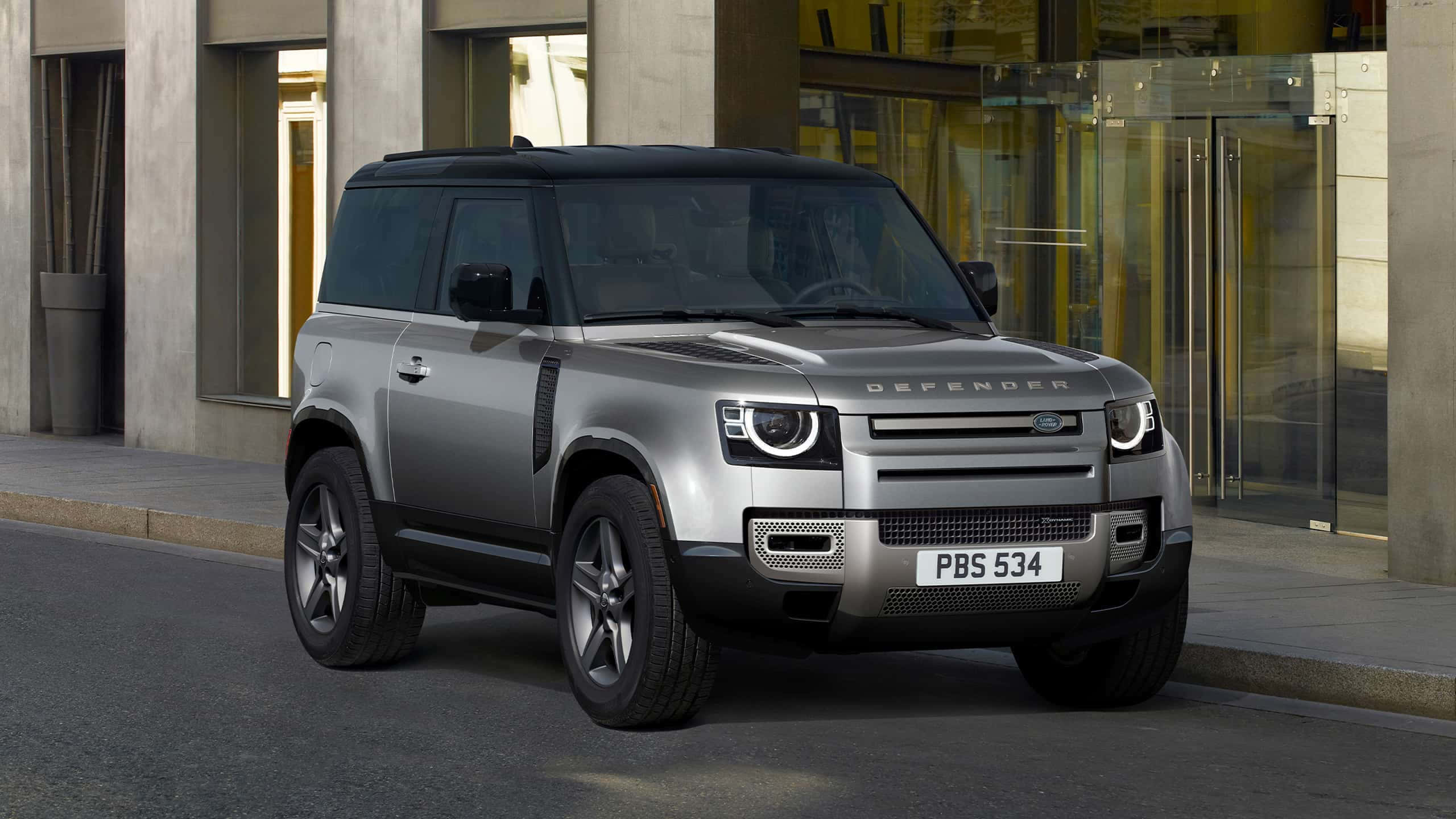 Land Rover Defender 桌布 壁紙 — 一輛時尚的銀色 Land Rover Defender 停泊在城市街道上，旁邊是一座現代玻璃建築。