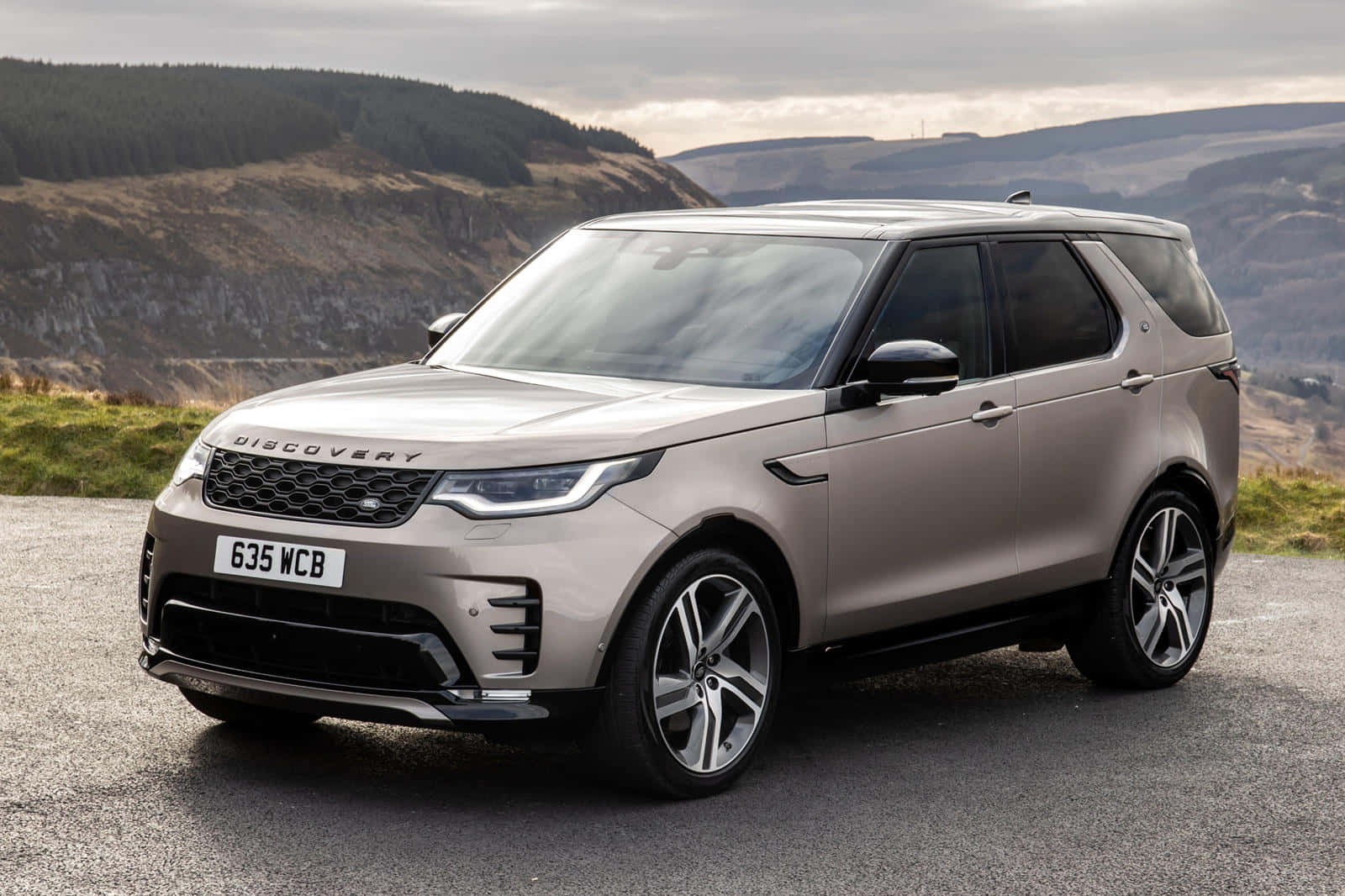 Land Rover Discovery 桌布 壁紙 — 一輛時尚的銀色Land Rover Discovery SUV停泊在碎石路上，背景是起伏的山丘和多雲的天空。