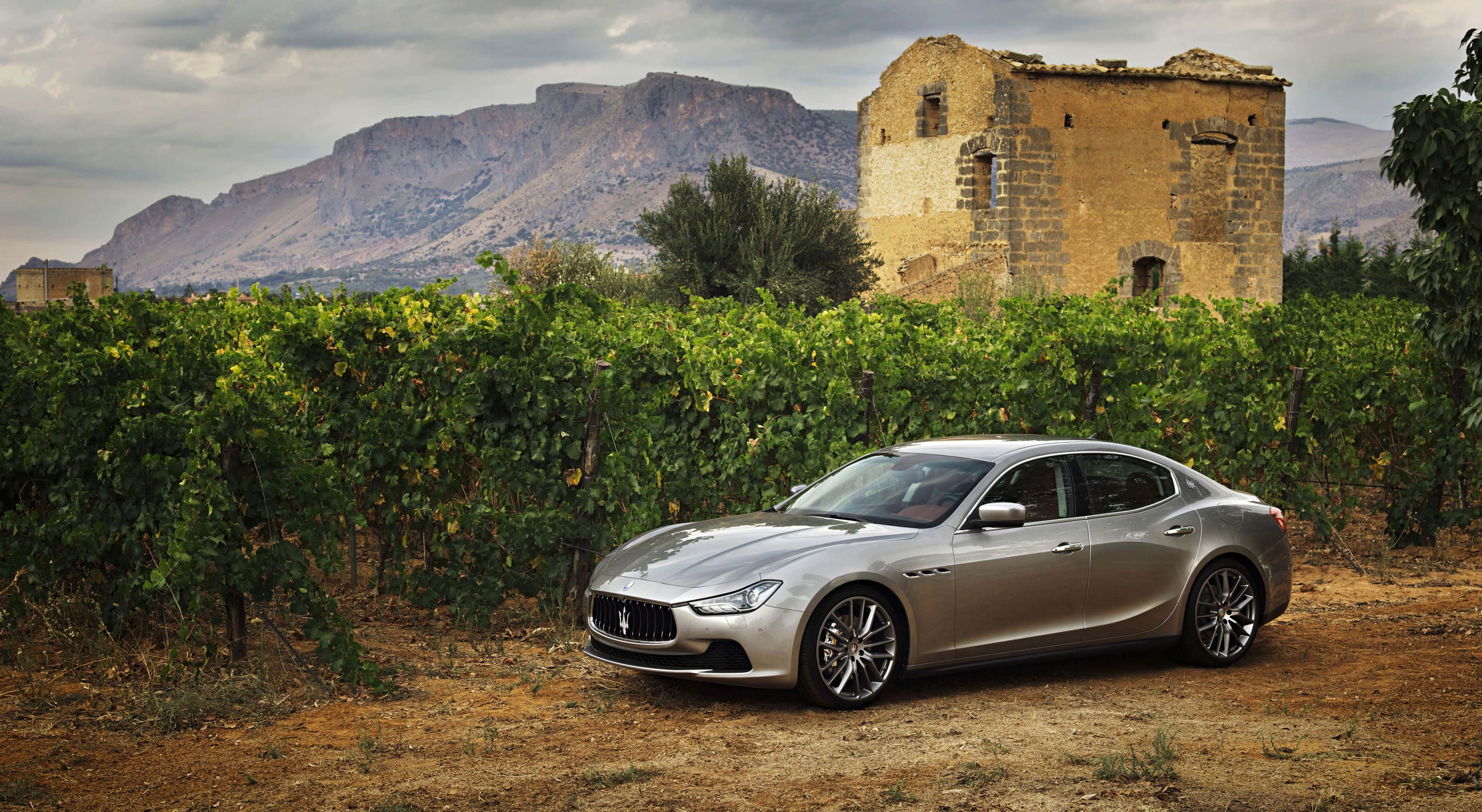 Maserati Ghibli 桌布 壁紙 — 一架時尚的銀色瑪莎拉蒂房車停泊在泥土上，旁邊是翠綠的葡萄園，背景有風化的石塔和崎嶇的山脈，天空多雲。