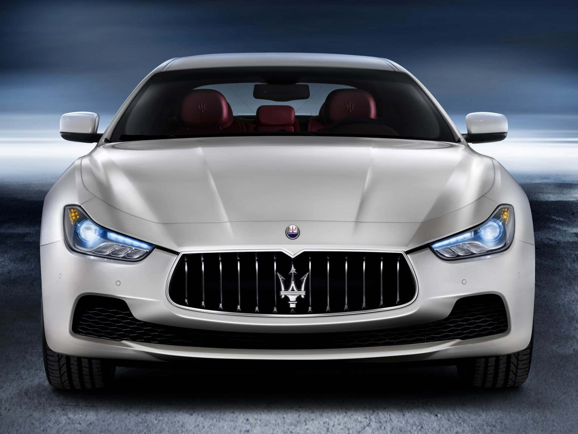 Maserati Ghibli 桌布 壁紙 — 一輛時尚的銀色瑪莎拉蒂跑車，配備發光頭燈和紅色皮革座椅，正面朝向戲劇性的深藍色背景。