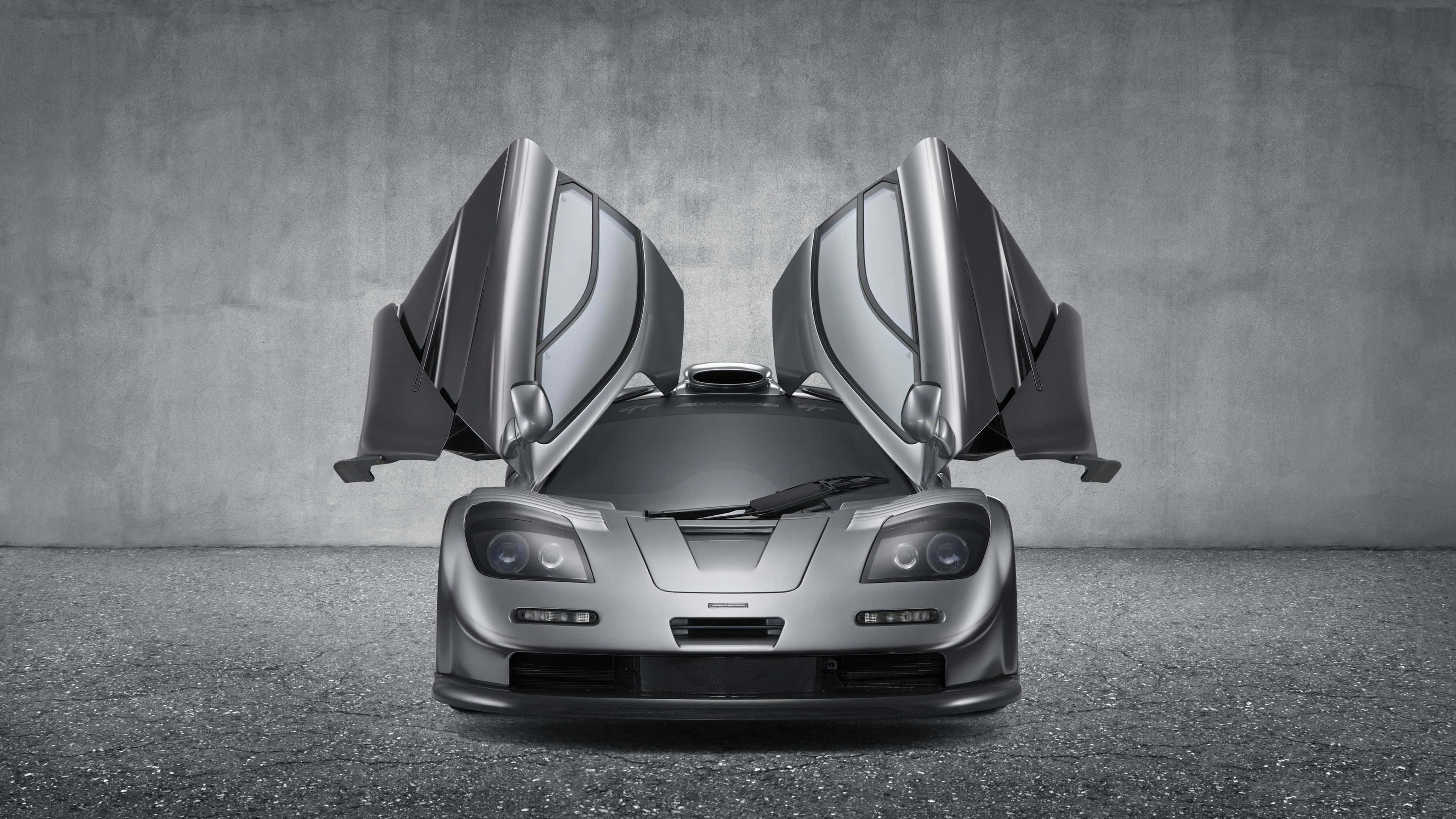 Mclaren F1 桌布 壁紙 — 一架流線型銀色McLaren F1超級跑車，鷗翼車門打開，停在簡約的混凝土牆前。
